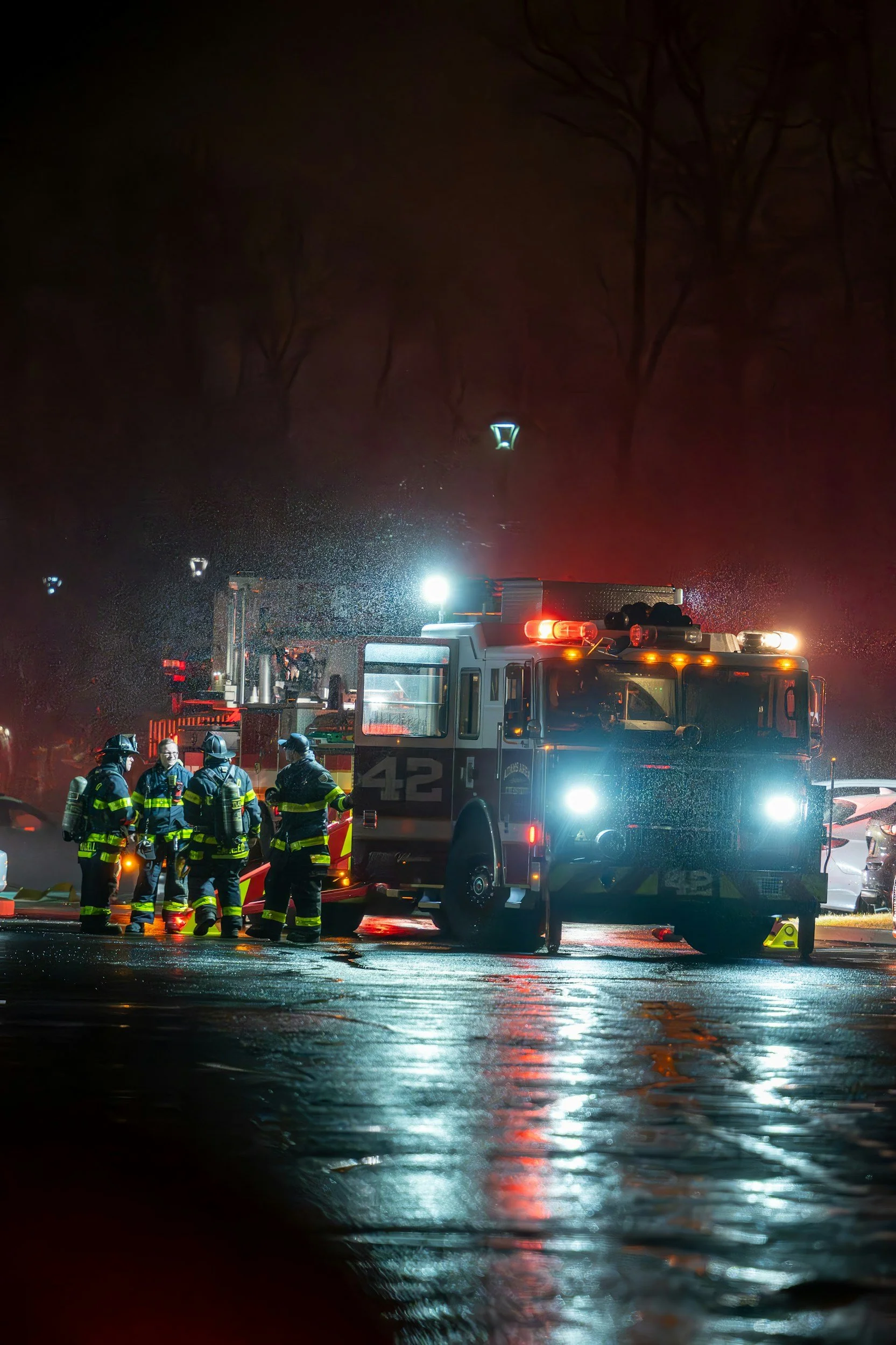 Firefighters in gear working at night next to a fire truck with lights flashing, wet pavement reflecting lights, and dark trees in the background.