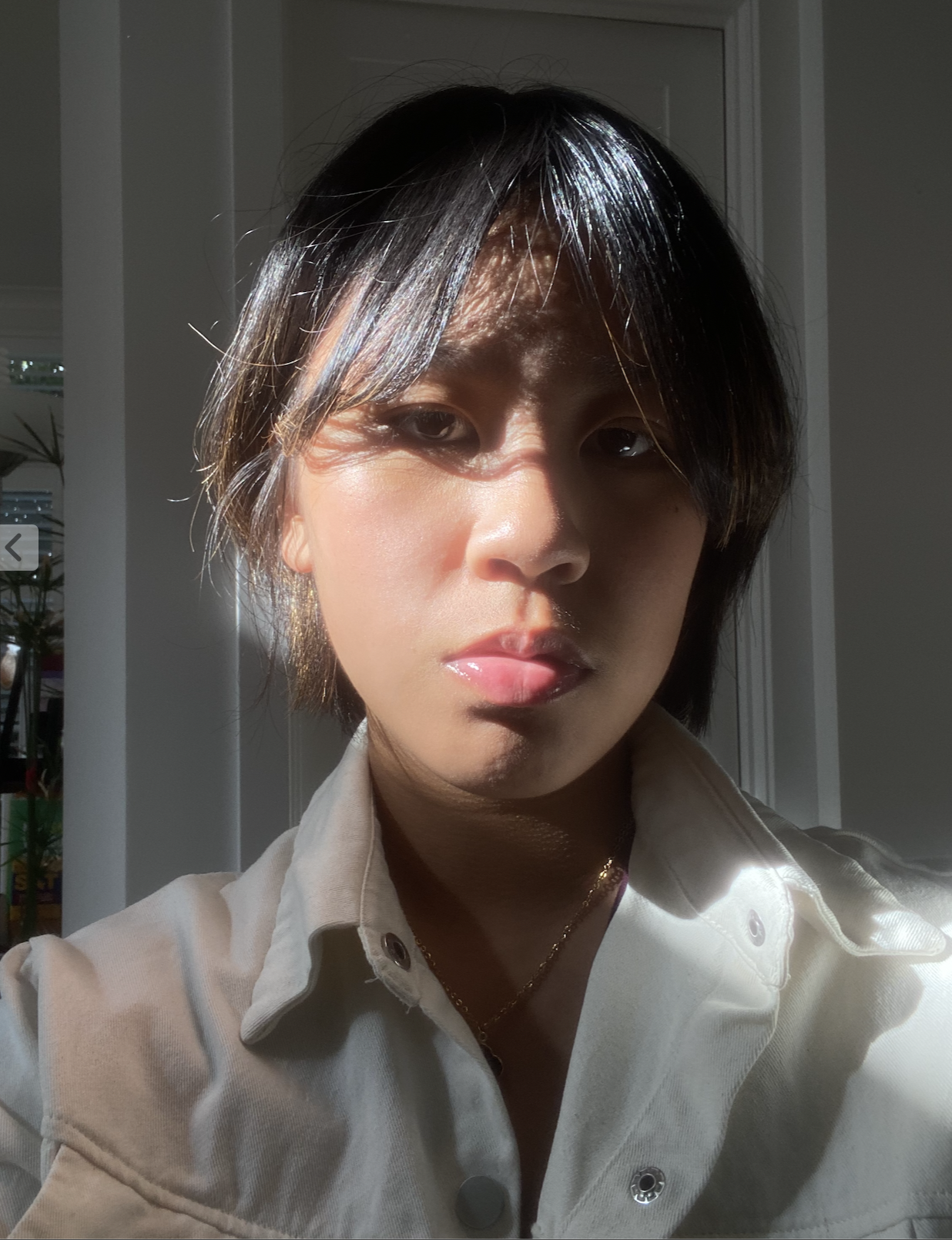 Person in partial sunlight with a neutral expression, wearing a light-colored collared shirt and a necklace.