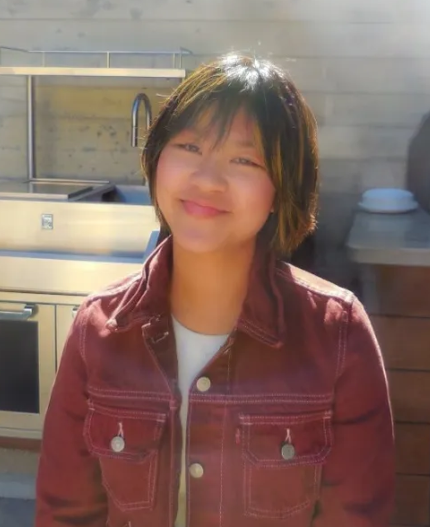 Person in partial sunlight with a neutral expression, wearing a light-colored collared shirt and a necklace.