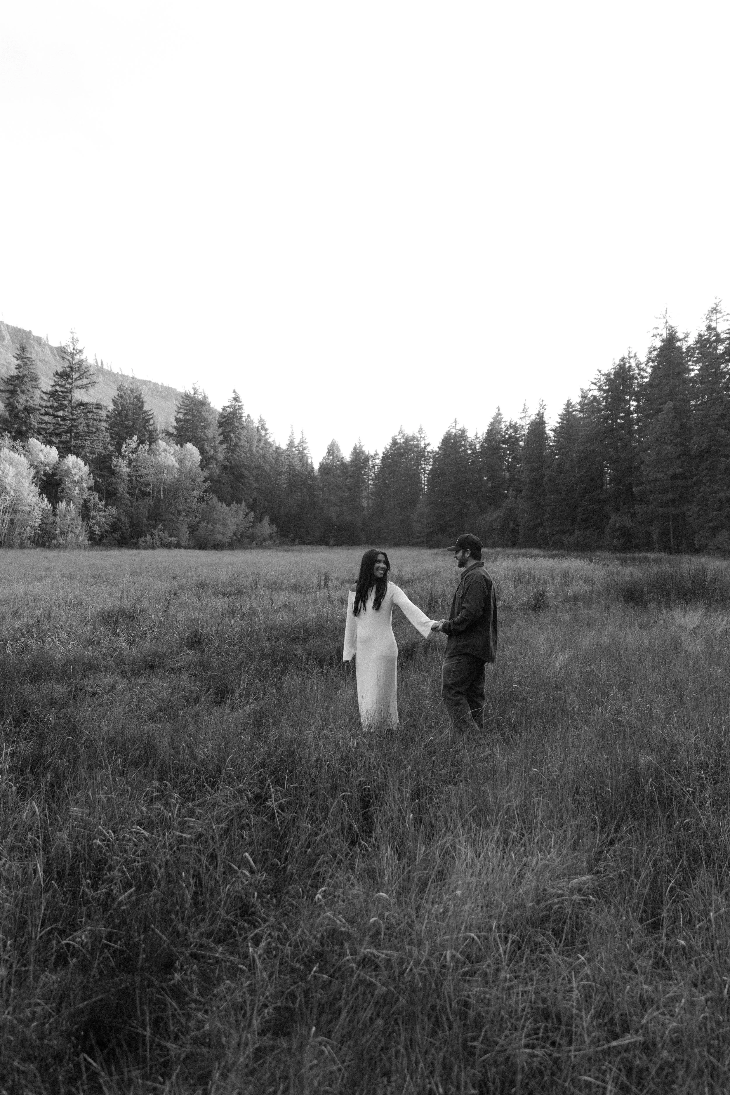 Black and white photo of a couple standing in a grassy field holding hands, surrounded by trees with mountains in the background.