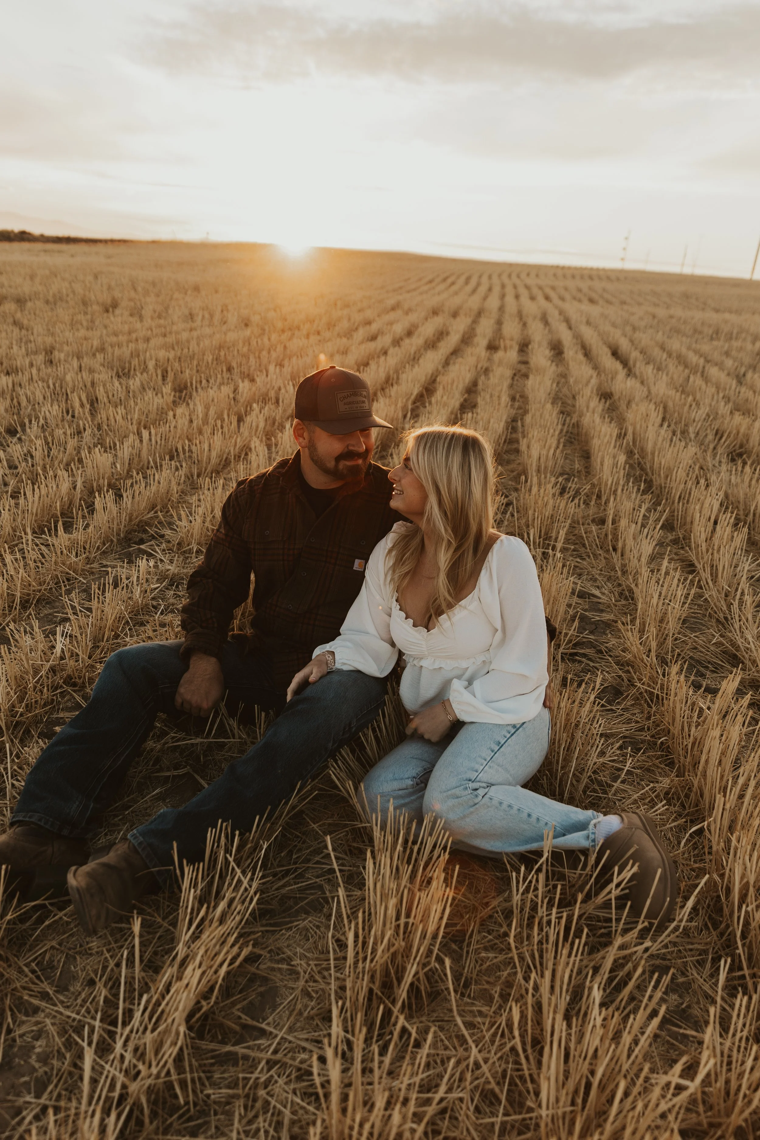A man and woman sitting on the ground in a harvested wheat field during sunset, looking at each other and smiling.