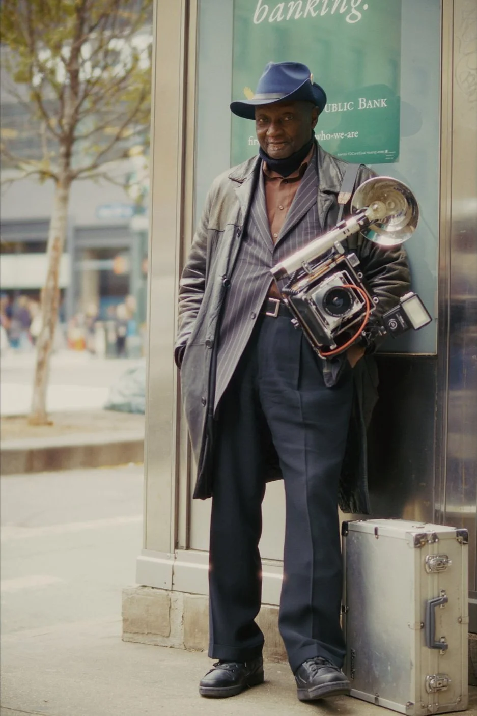 A man in a black hat and leather coat stands on a city sidewalk, holding a vintage camera with a horn attached, next to a silver case, while people walk by in the background.