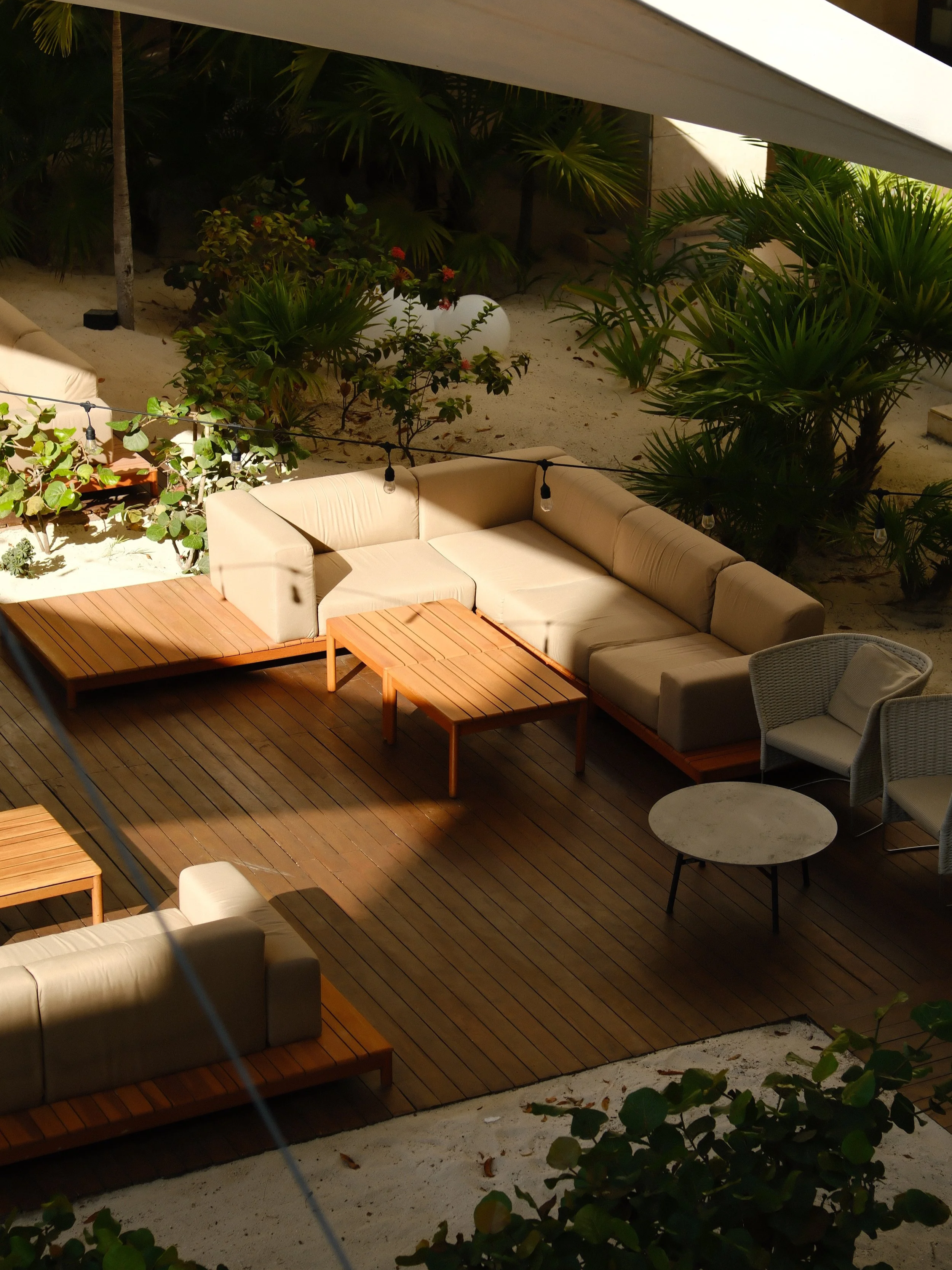 An outdoor patio with beige cushioned sofas and wooden tables, surrounded by green tropical plants and sand, illuminated by hanging string lights.