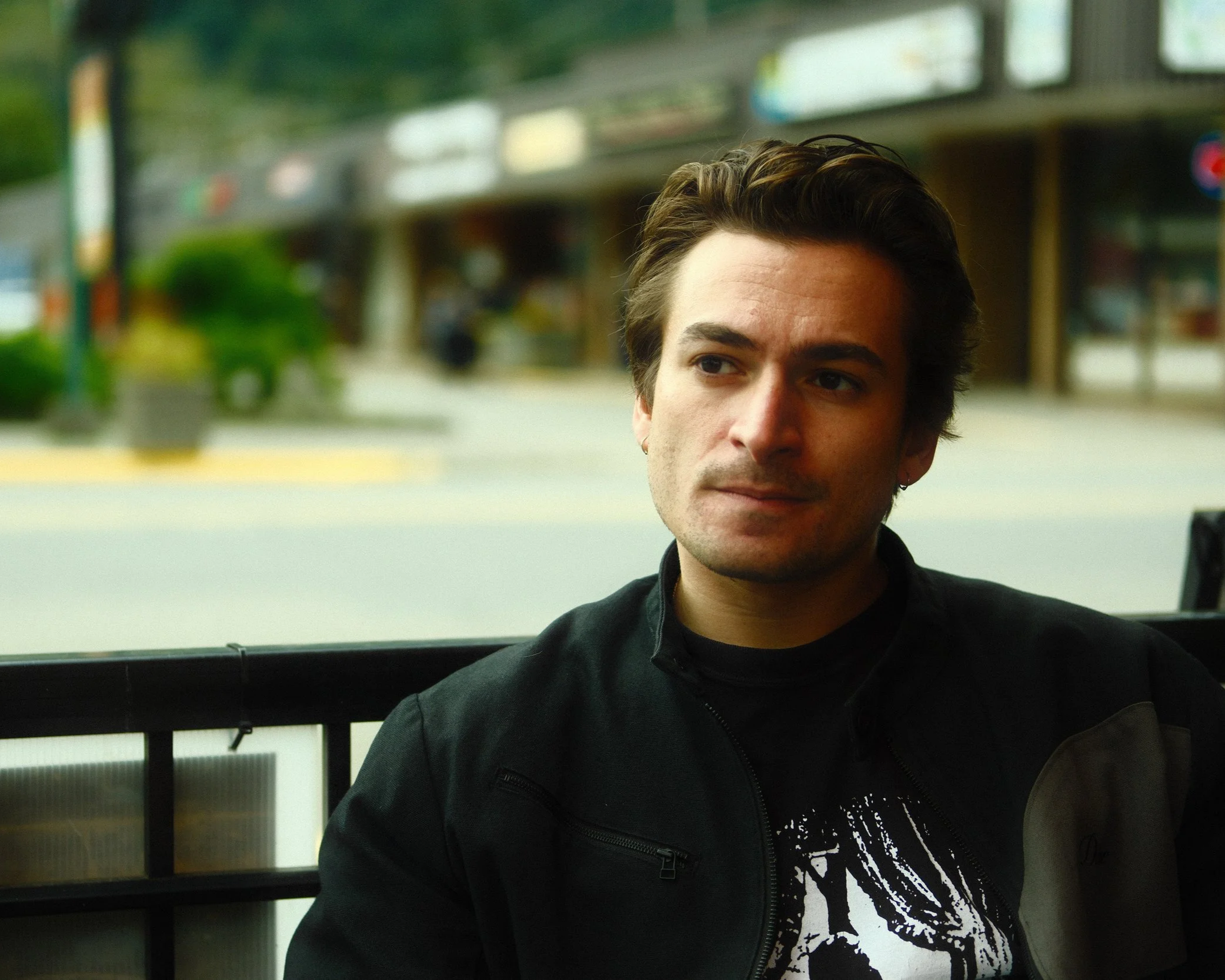 A man with dark hair and a black jacket sitting outdoors, with a city street background.