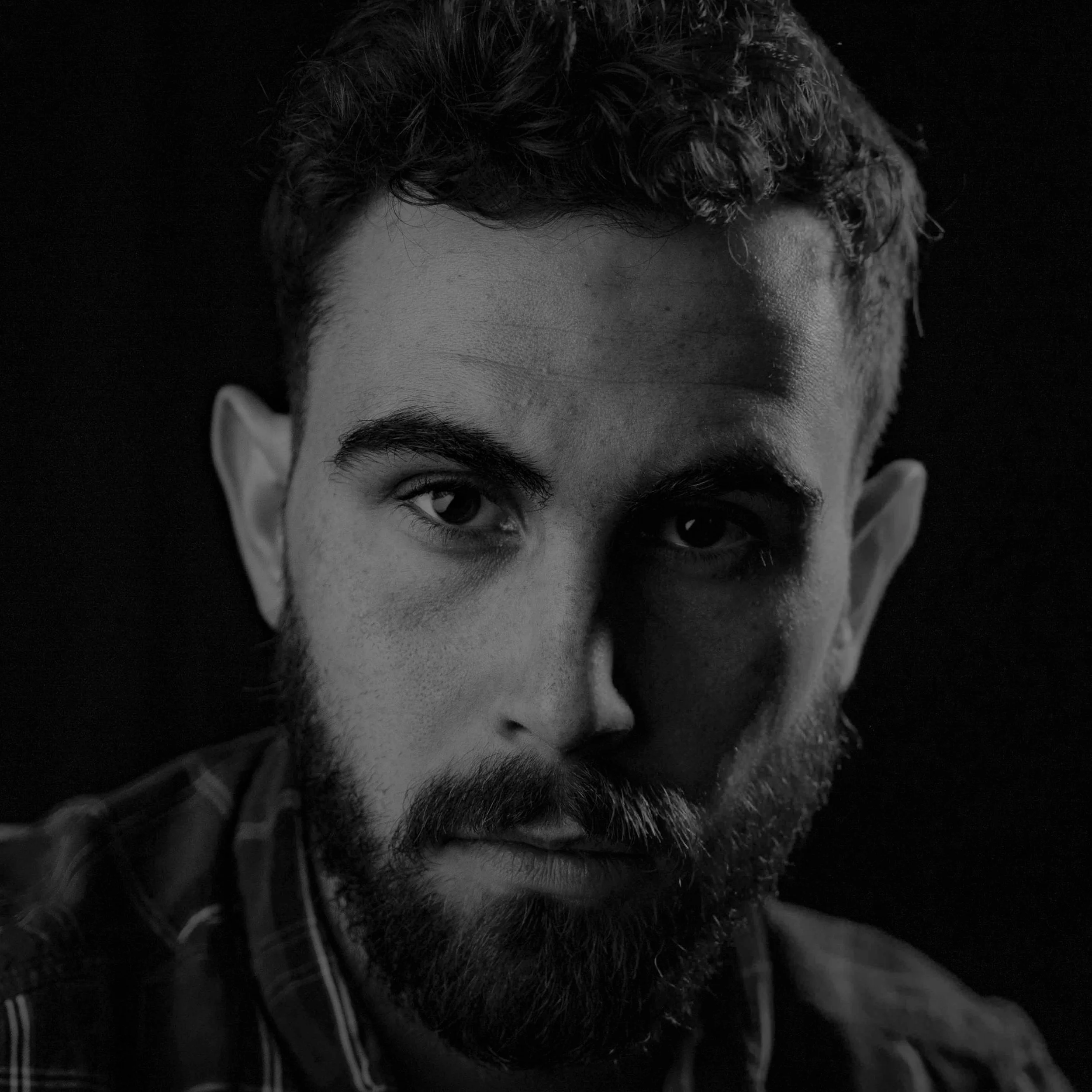 Black and white portrait of a bearded man with short curly hair, looking directly at the camera.
