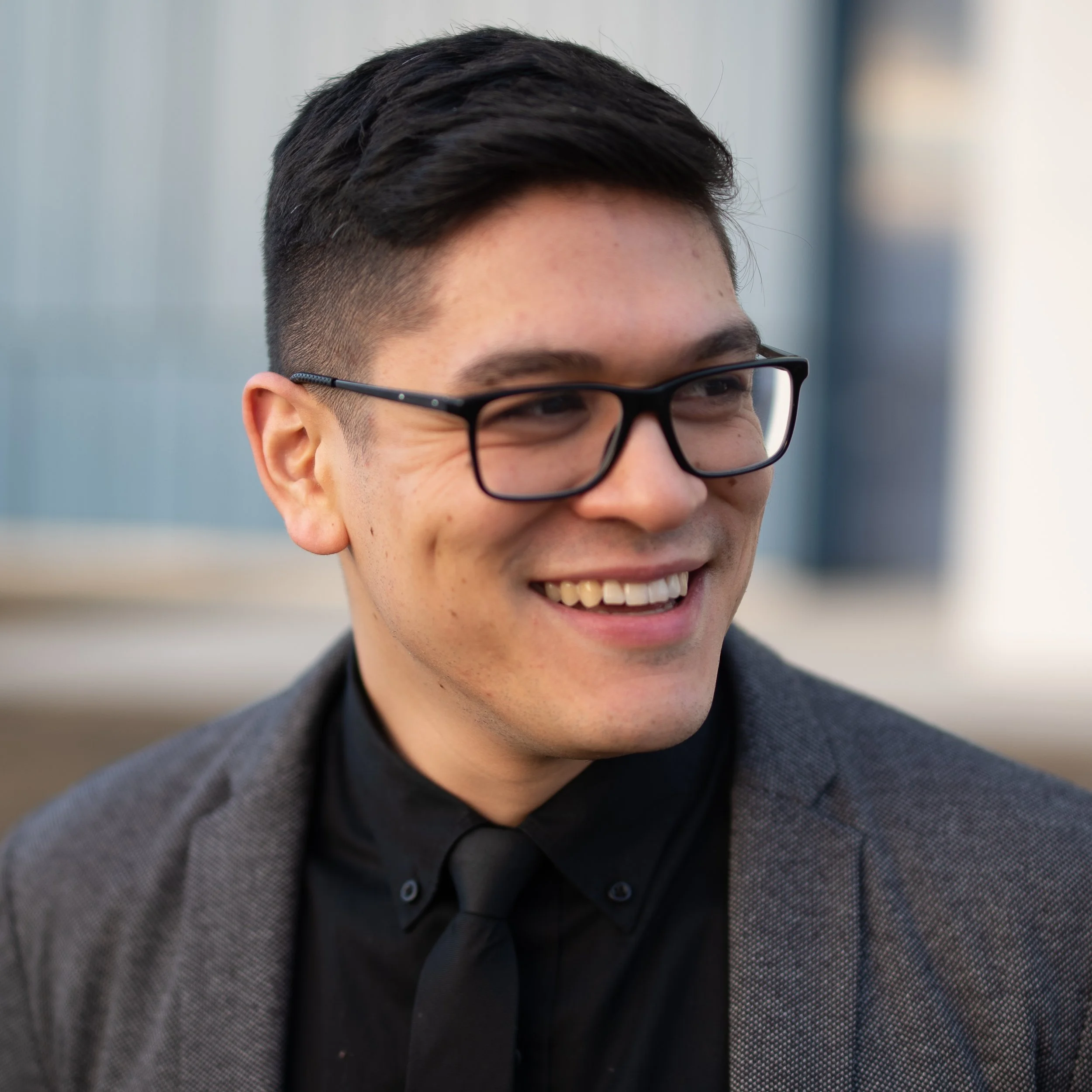 Smiling person wearing glasses and a suit jacket with a black shirt and tie, looking to the side.