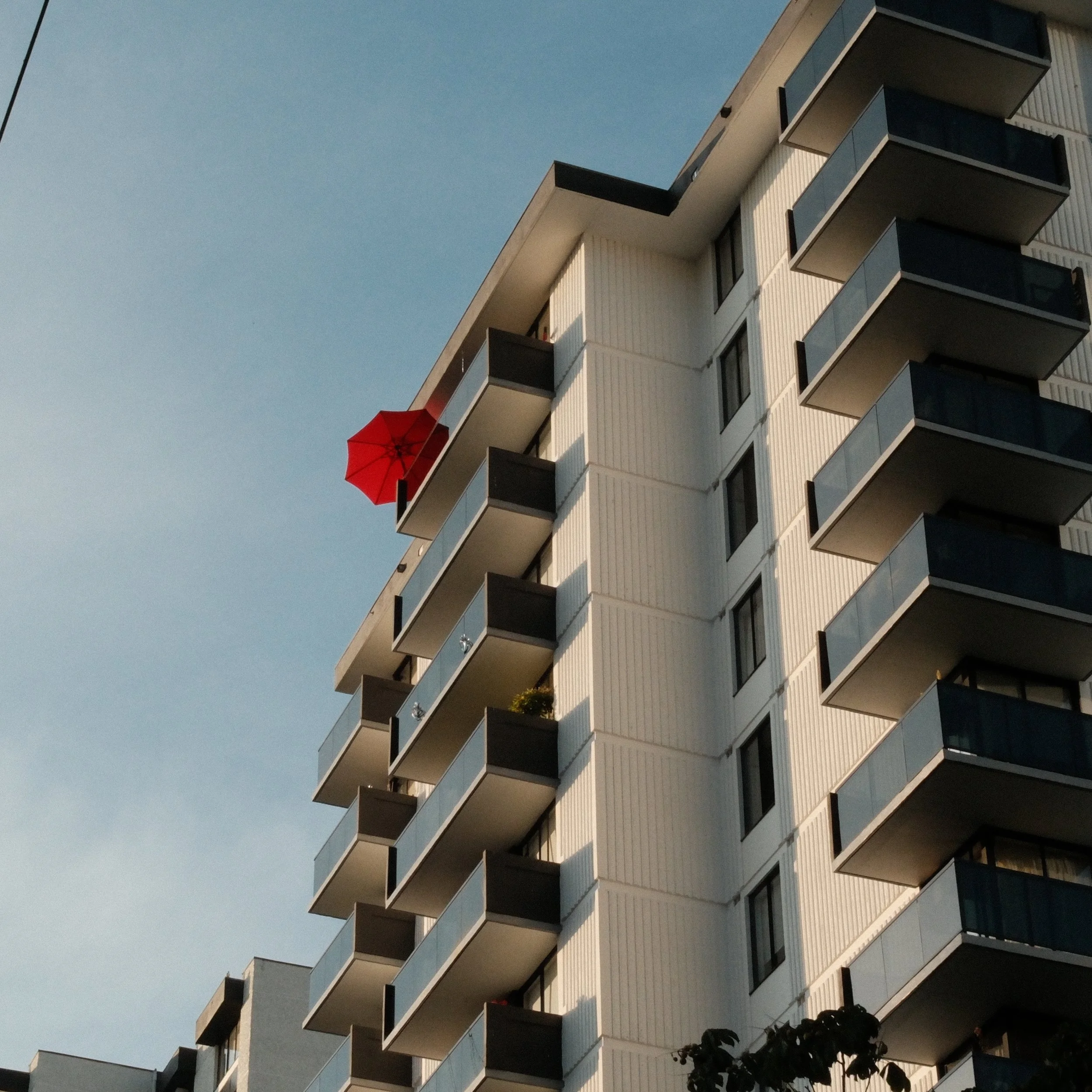A multi-story apartment building with balconies, one of which has a red umbrella, against a blue sky.