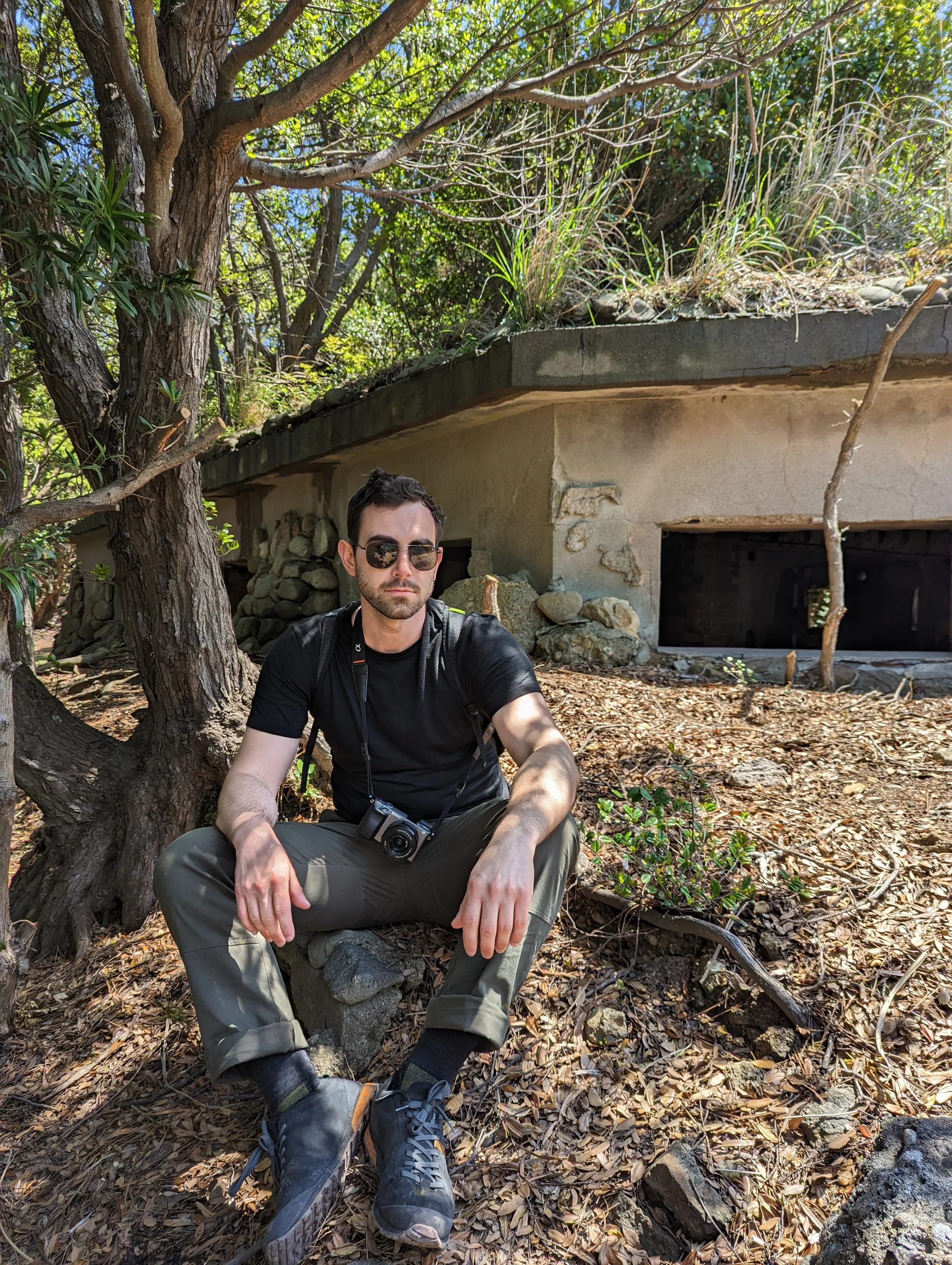 A man with sunglasses sitting on a rocky ground in a wooded area with trees and a small structure in the background.