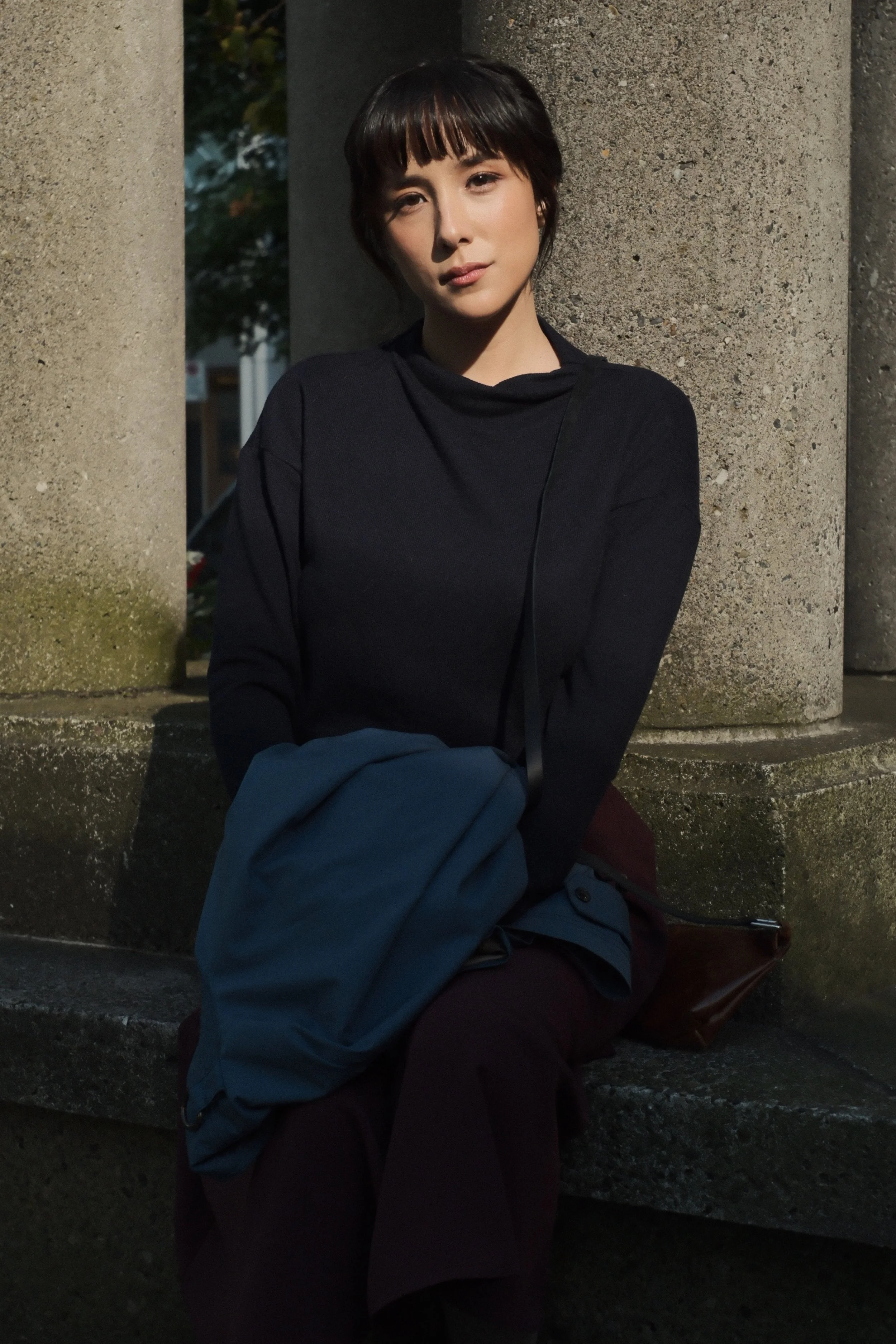 A woman with short dark hair and bangs, wearing a black top, sitting outdoors against stone columns, holding a blue jacket and looking at the camera.