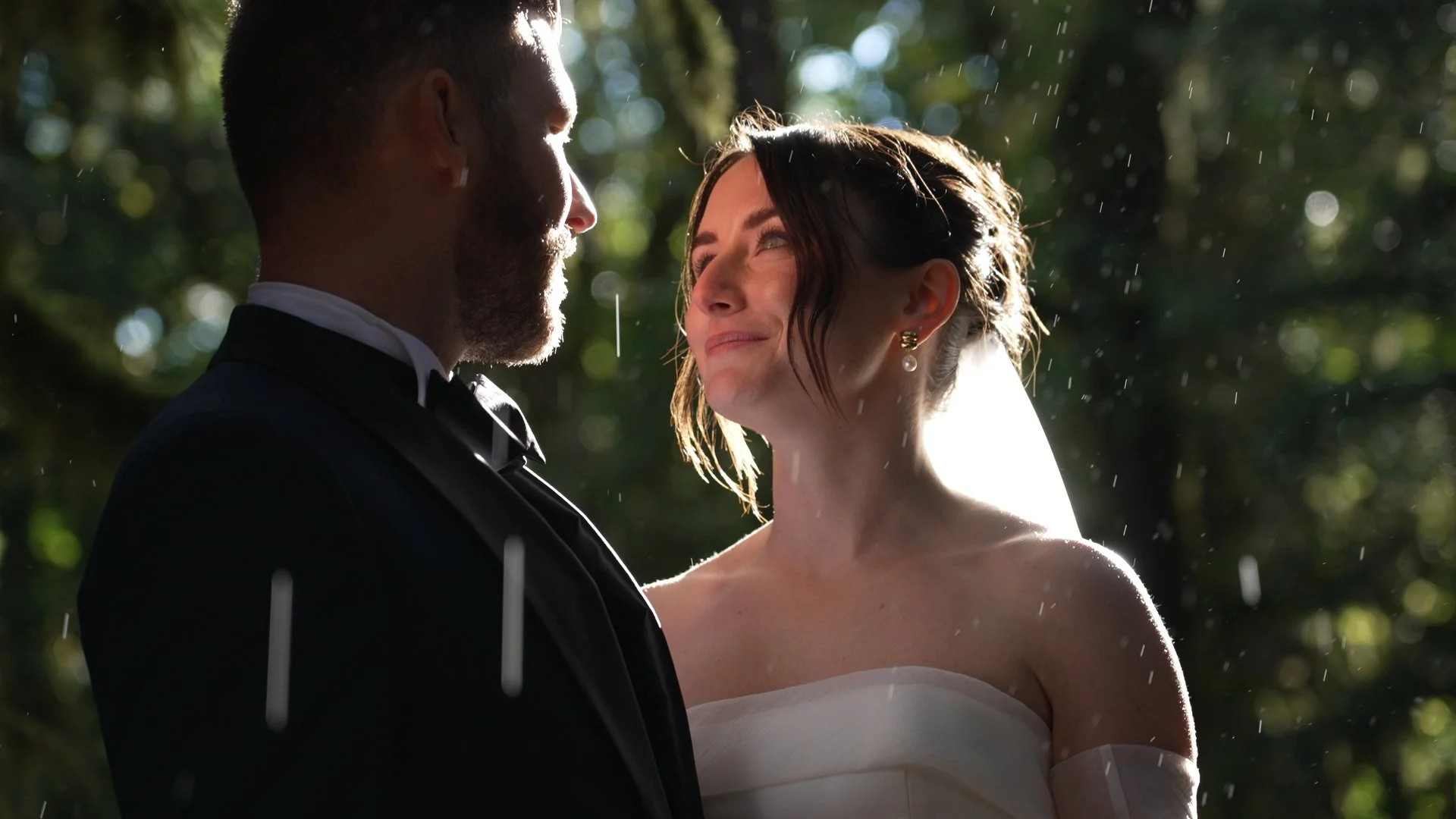 A bride and groom standing close together outdoors in the rain, looking into each other's eyes, with sunlight filtering through trees in the background.