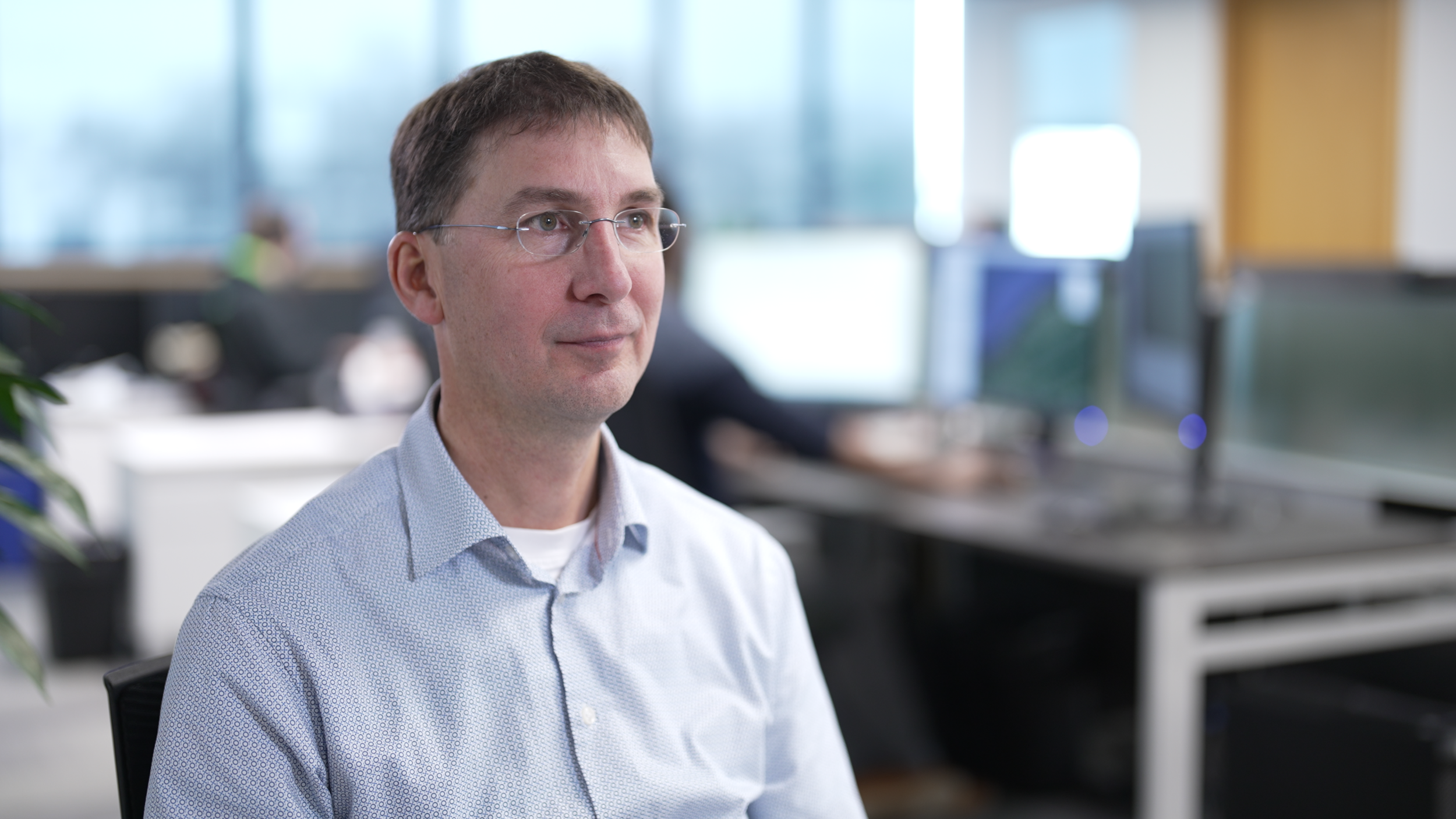 A man with glasses wearing a light blue shirt sitting in an office with computer monitors in the background.