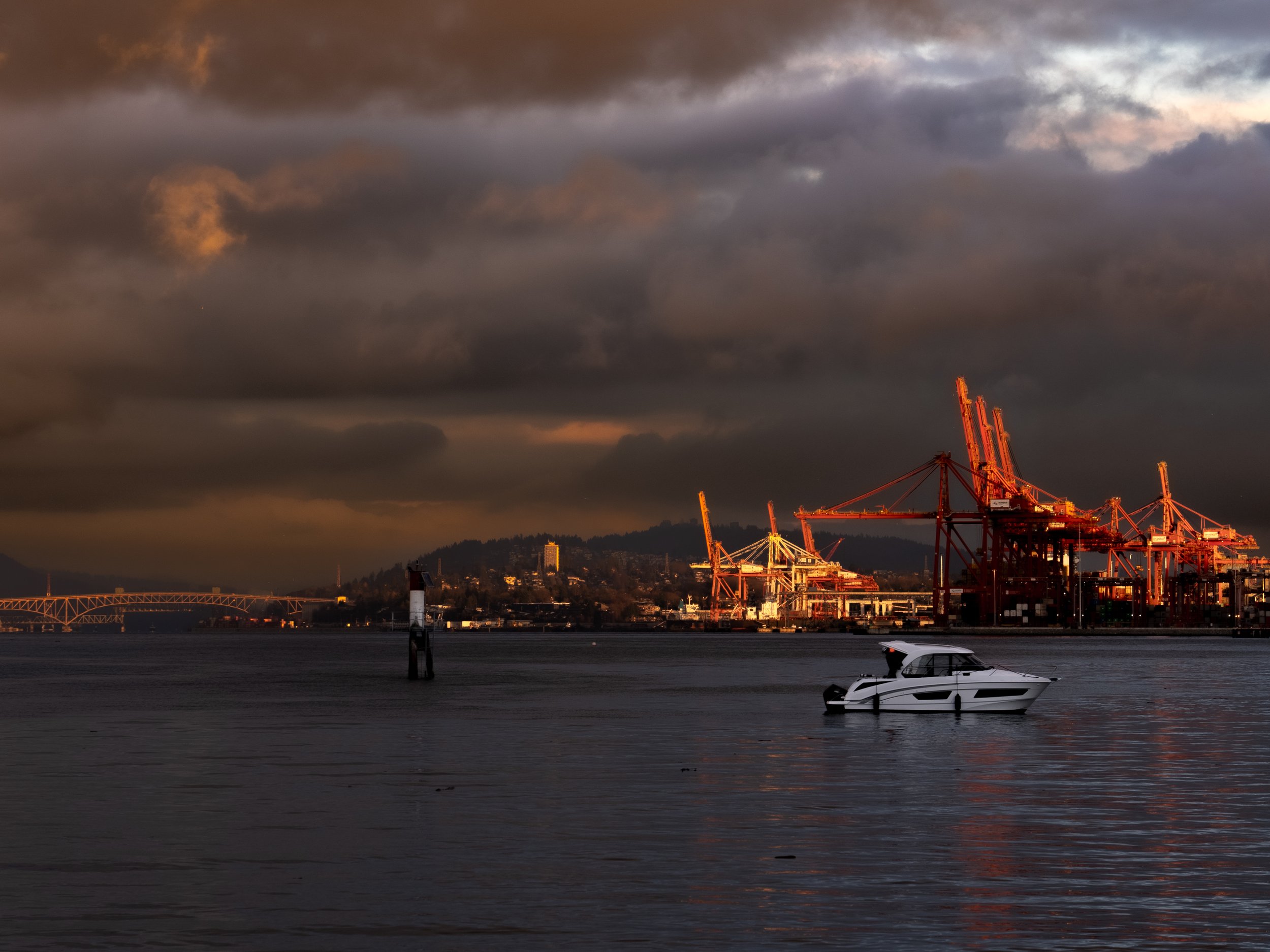 A boat on calm water with a dark, cloudy sky and an industrial port with cranes in the background during sunset.