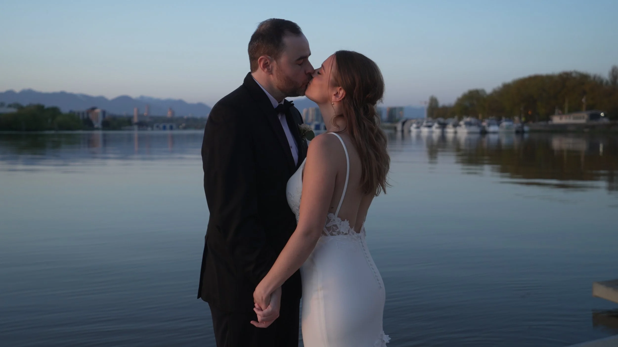 A newlywed couple kissing by a calm waterfront with boats and trees in the background at sunset