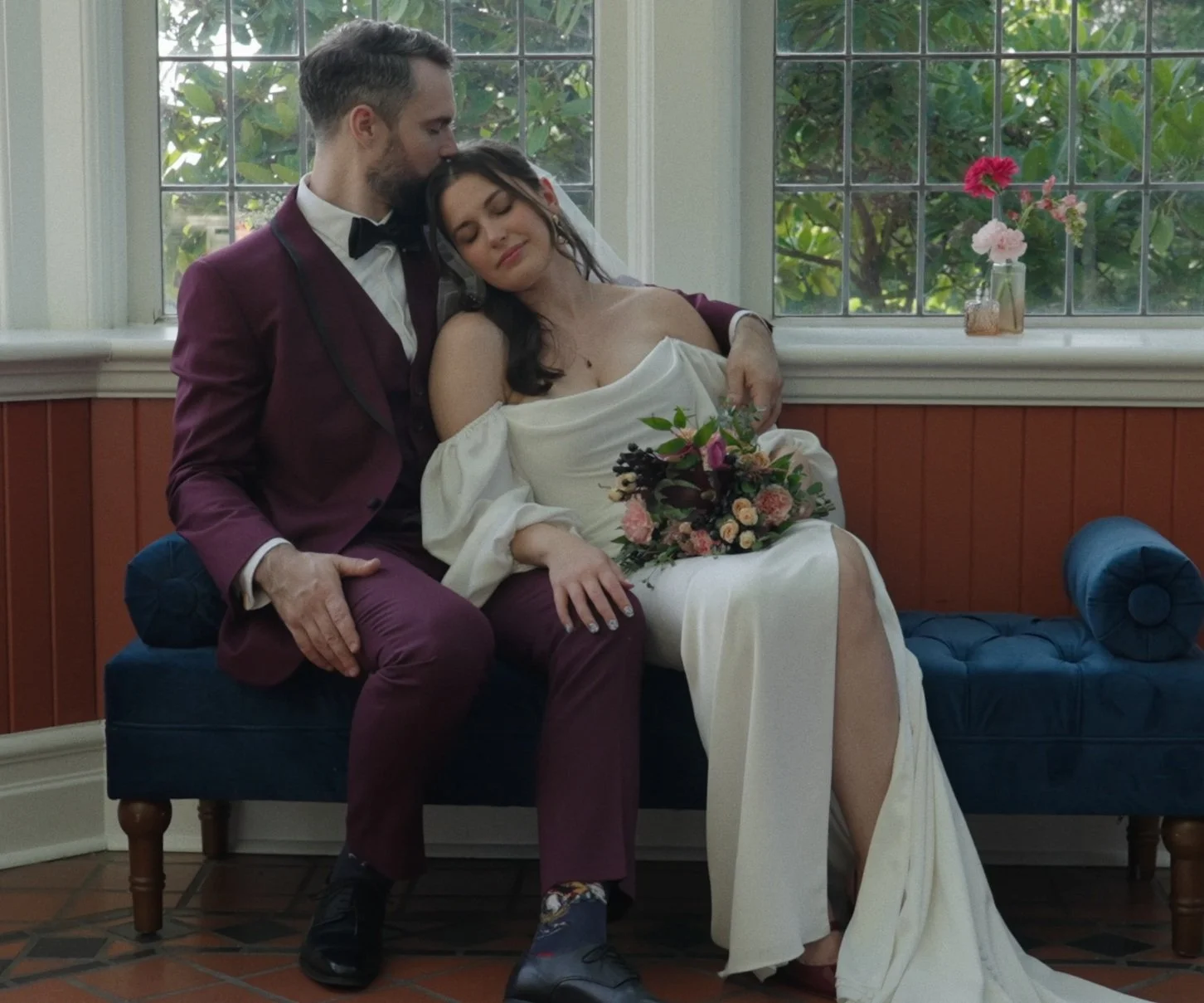 A couple sitting on a blue bench in a room with large windows and wooden paneling, with the man giving a gentle kiss on the woman's forehead. The woman is in a white wedding dress holding a bouquet of flowers, and the man is in a dark red suit