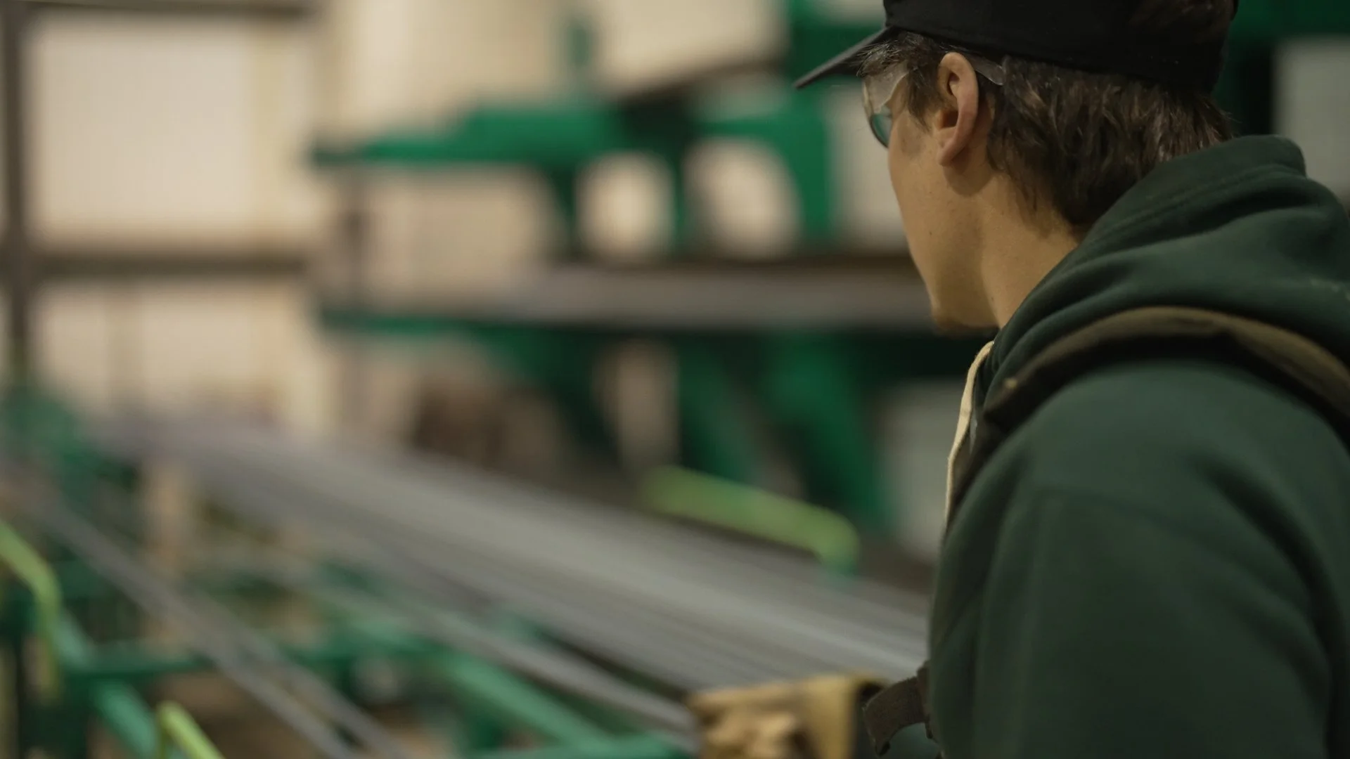 A young man in a green hoodie and a black cap is working in a workshop or industrial setting, looking away from the camera, with shelves and equipment in the background.