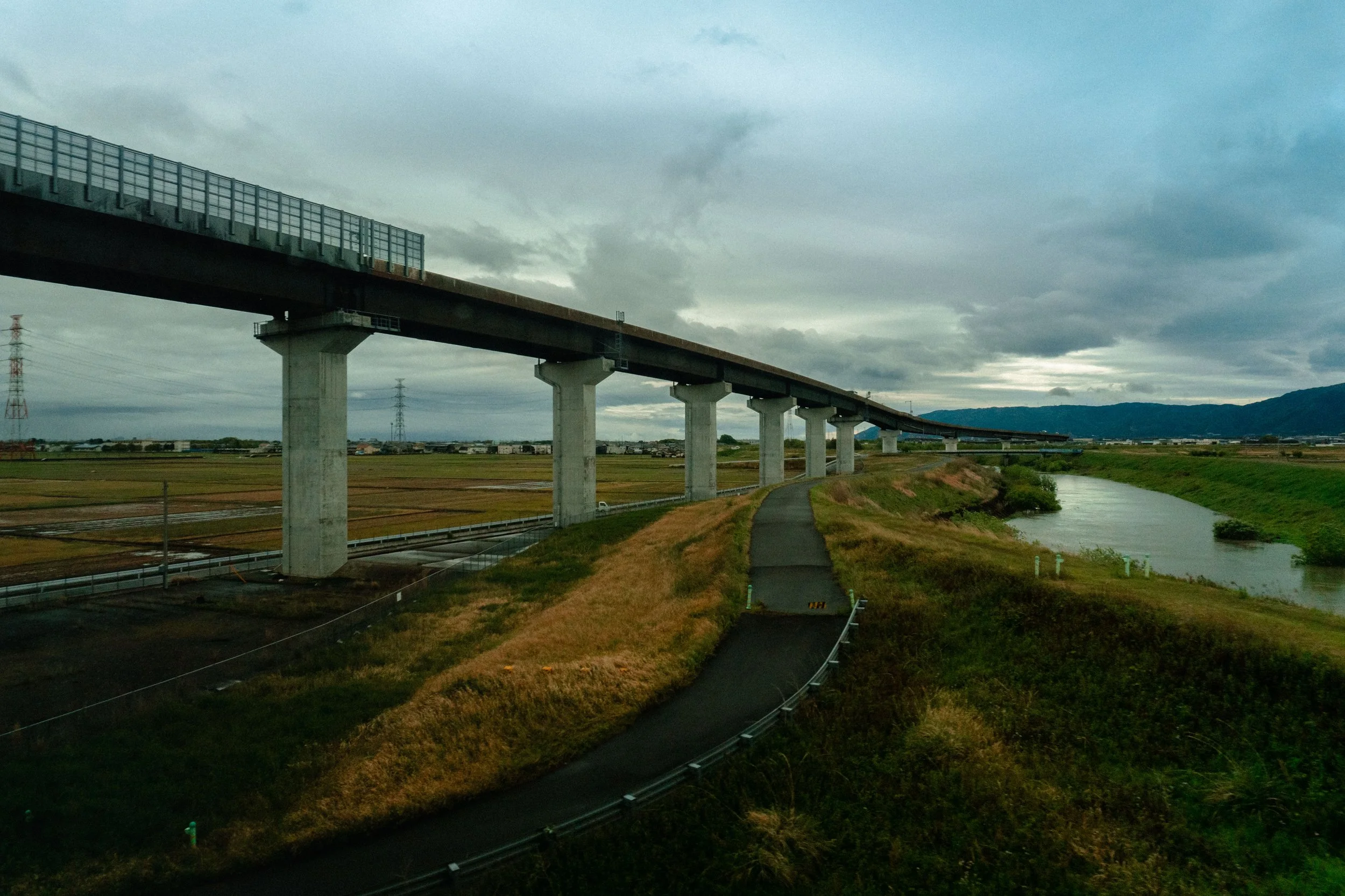 A long elevated highway extends over a rural landscape with a narrow waterway running beside it, under a cloudy sky.
