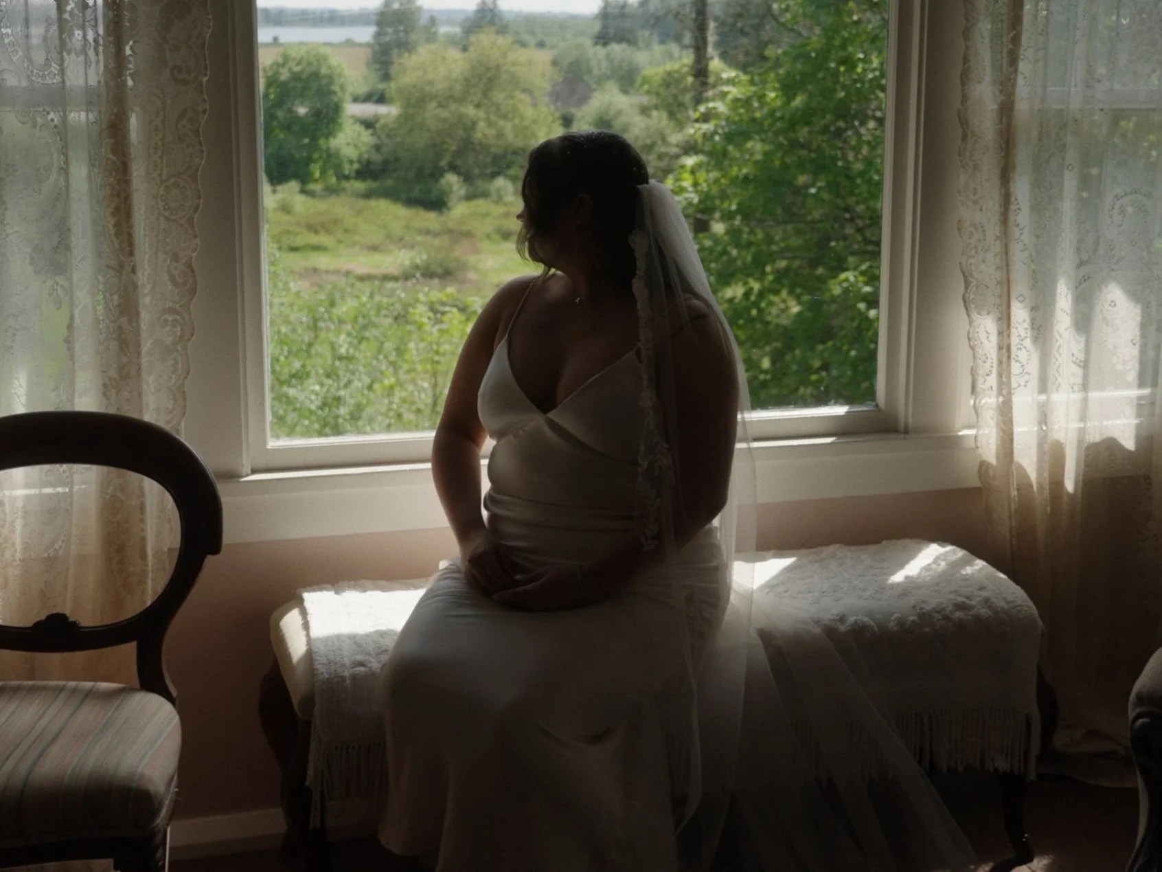 Silhouette of a woman wearing a wedding dress and veil, sitting on a bench by a window with lace curtains, overlooking green trees and a field.
