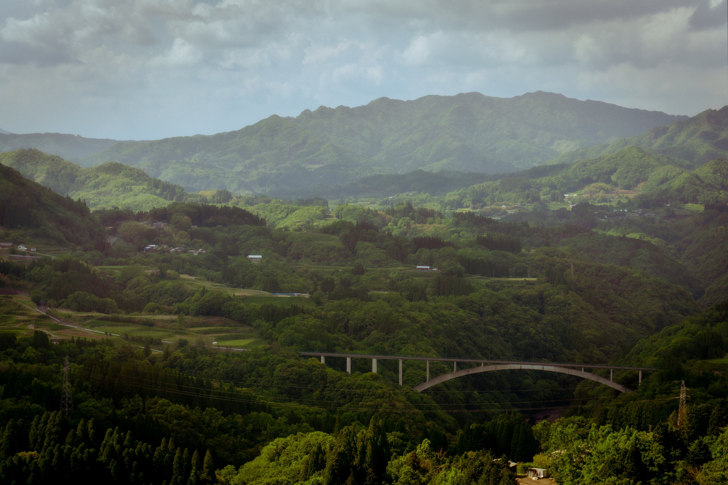 Scenic view of a lush, green landscape with rolling hills and a bridge in the foreground. Mountains are visible in the background under a partly cloudy sky.