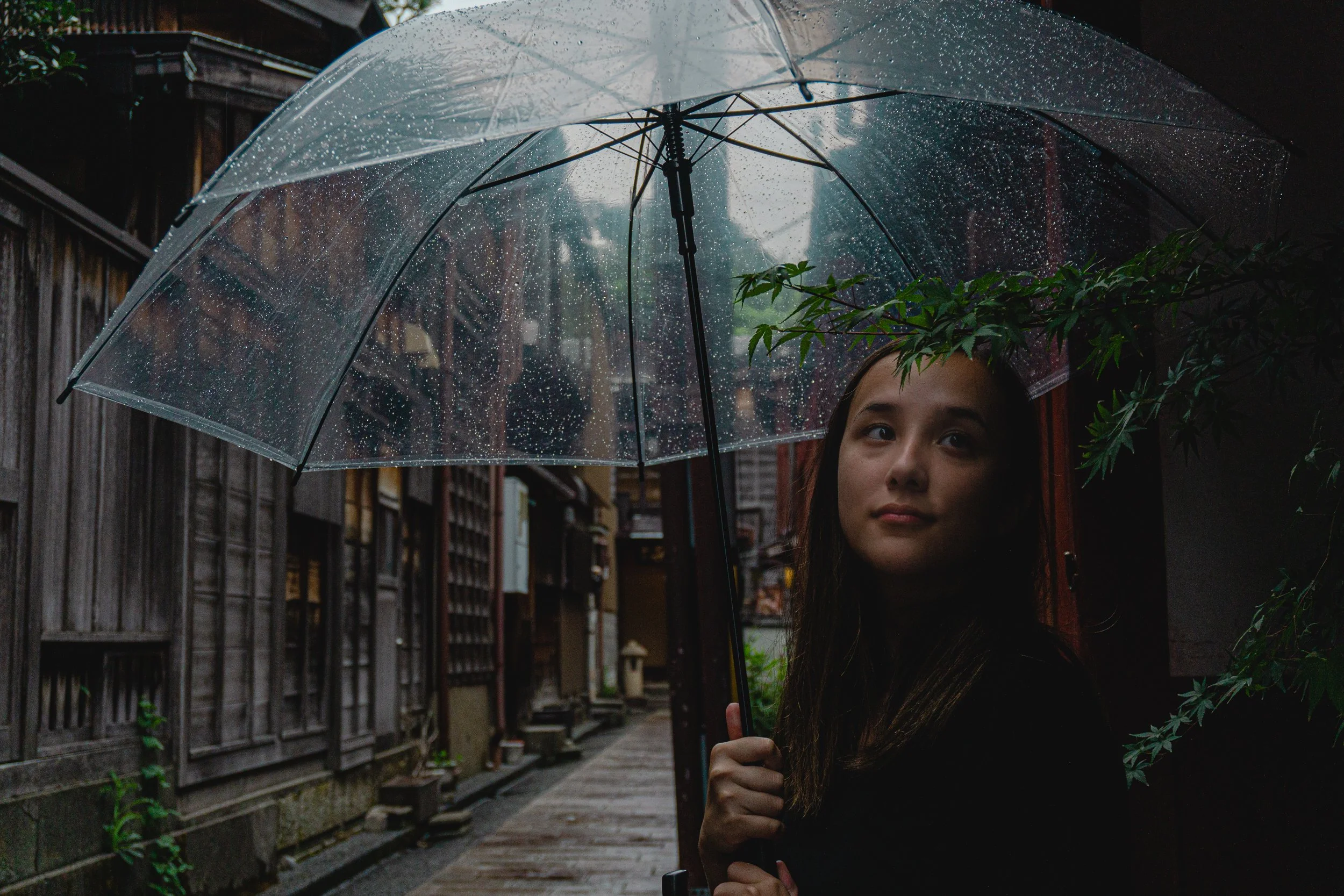 A woman holding a transparent umbrella with raindrops, standing in an alley with traditional wooden buildings and green foliage.