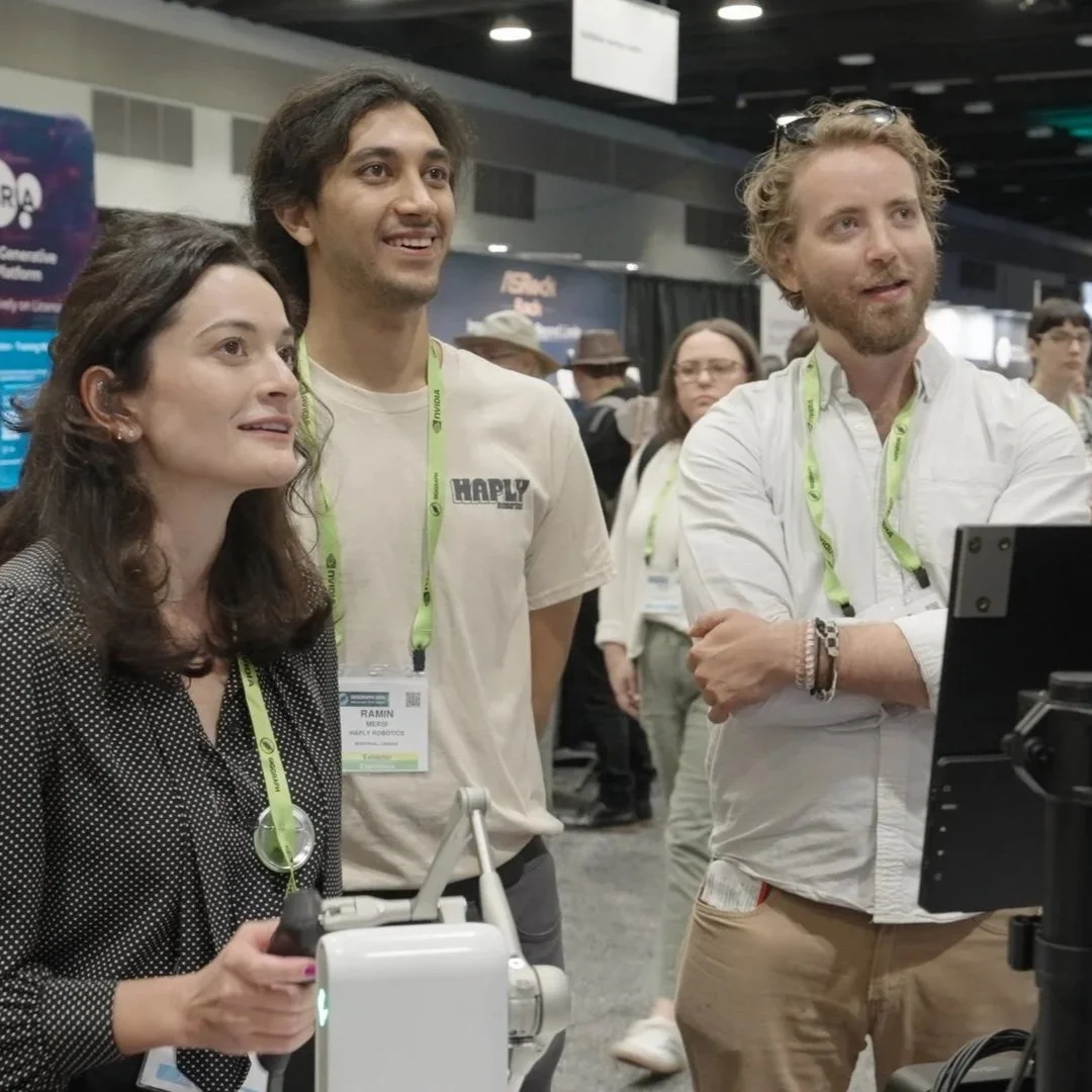 Two men in safety glasses and dark shirts are having a discussion in an industrial or manufacturing setting, with one man gesturing with his hand.
