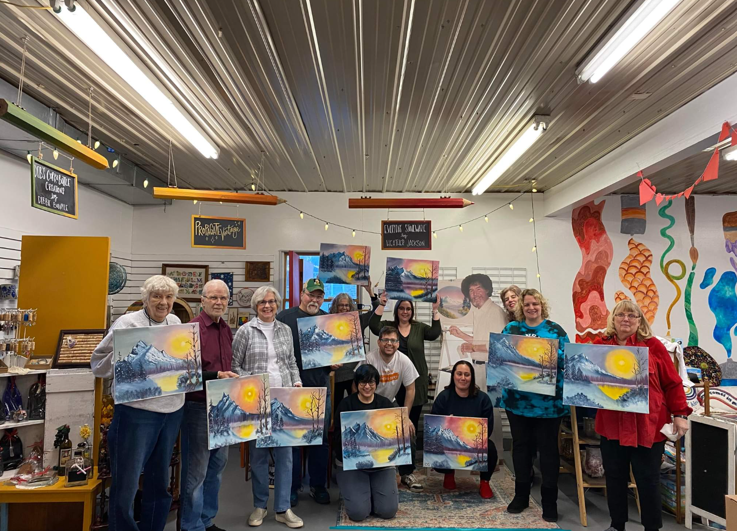 Group of people in an art studio holding finished landscape paintings of a sunset over mountains, with art supplies and colorful wall murals in the background.