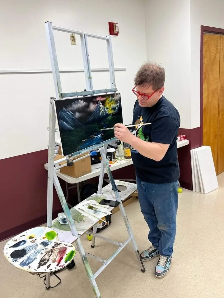 Person painting landscape on easel with art supplies in room