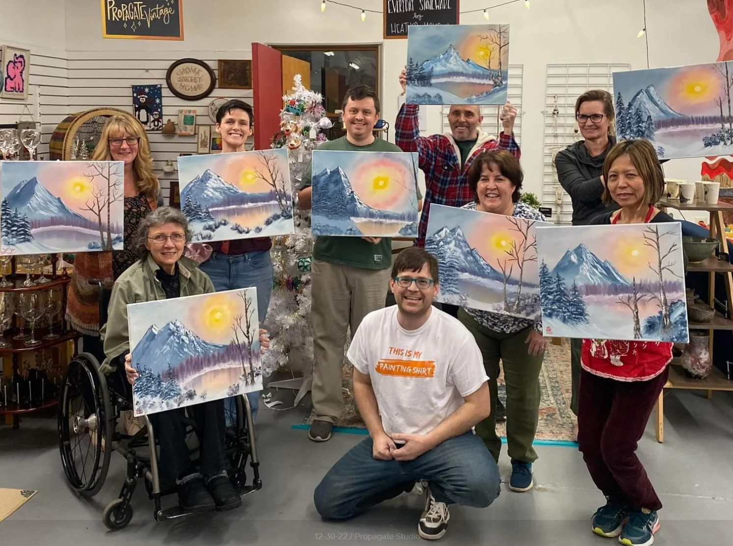 A group of people holding paintings of a winter mountain landscape scene inside an art studio.