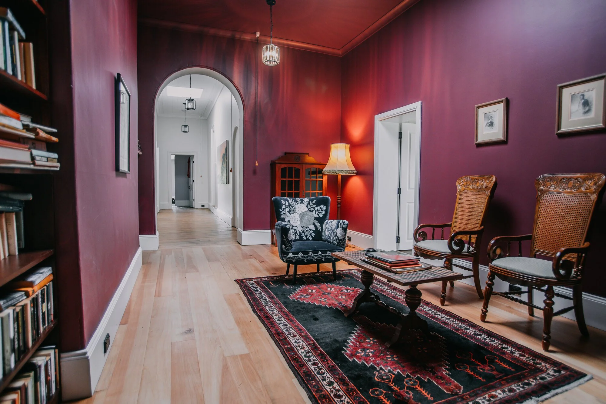 Living room with red walls, a patterned black and red rug, a blue armchair with floral embroidery, two wooden chairs, a wooden coffee table with magazines, a bookshelf on the left, a floor lamp, and wall art.