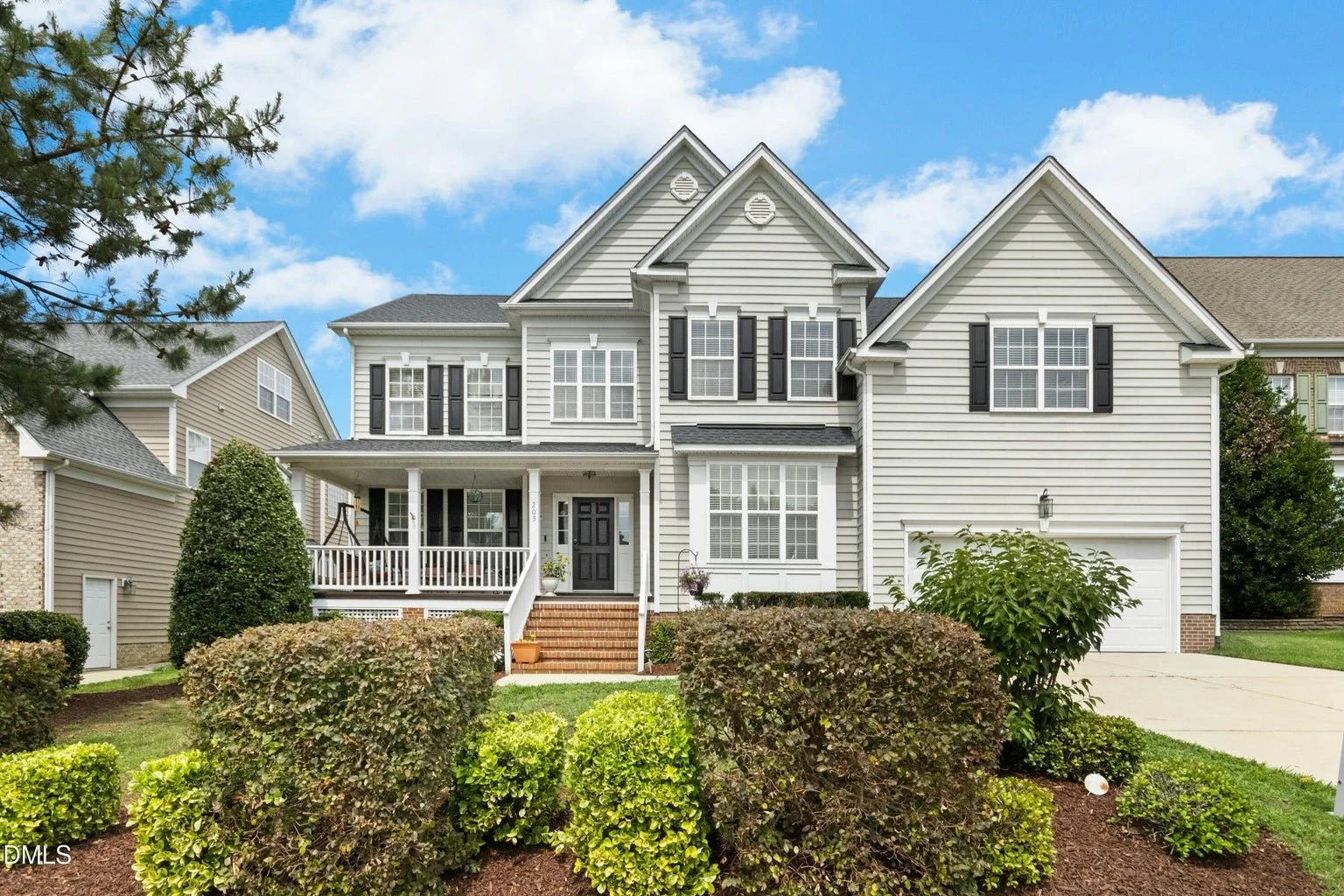 A large, two-story suburban house with white siding and black shutters, front yard with bushes and a small tree, brick steps leading to a covered porch with white railings, attached garage, and a bright blue sky with some clouds in the background.