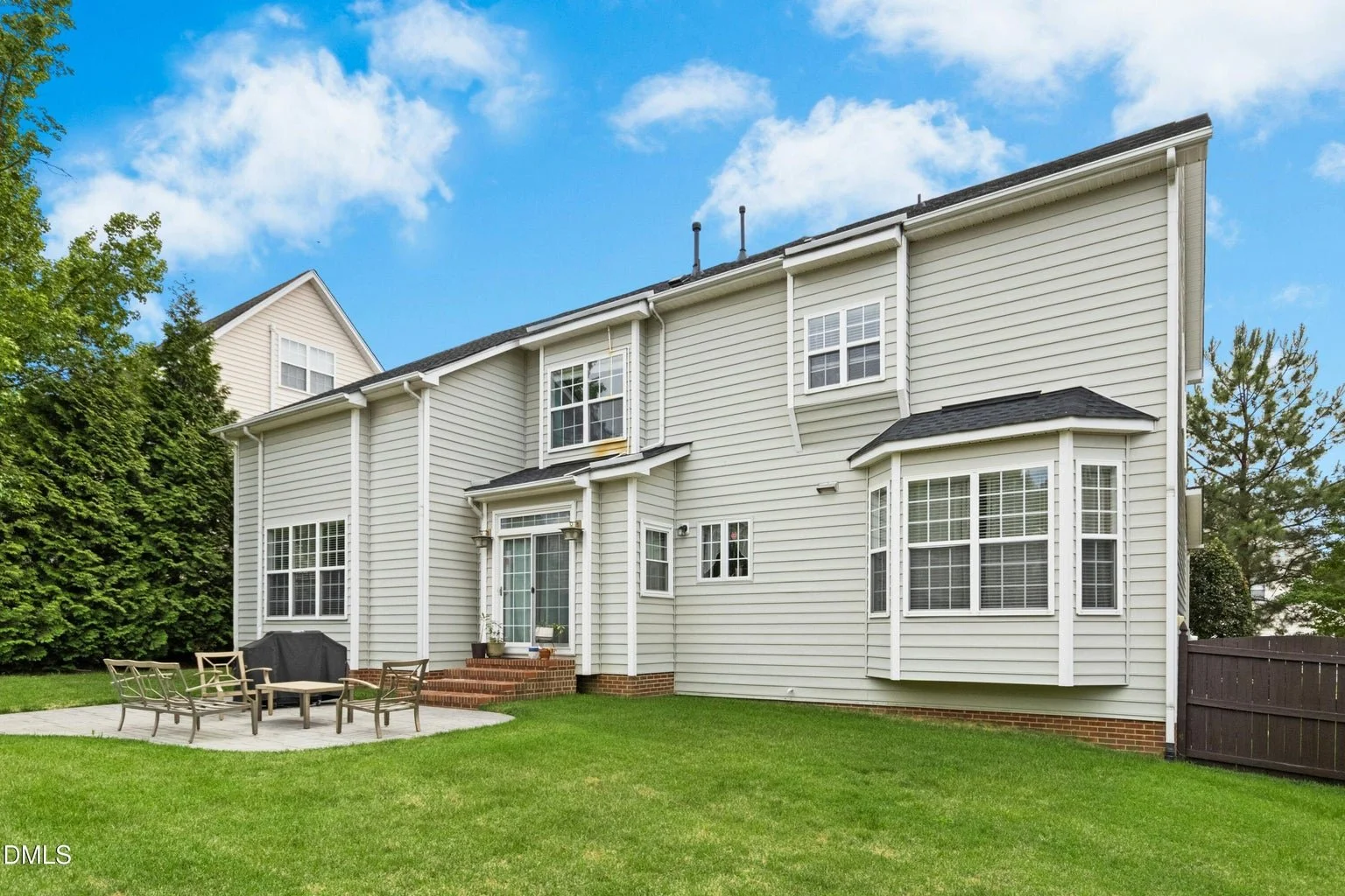 Backyard of a white two-story house with a small patio, outdoor furniture, a barbecue grill, and a green lawn under a partly cloudy sky.