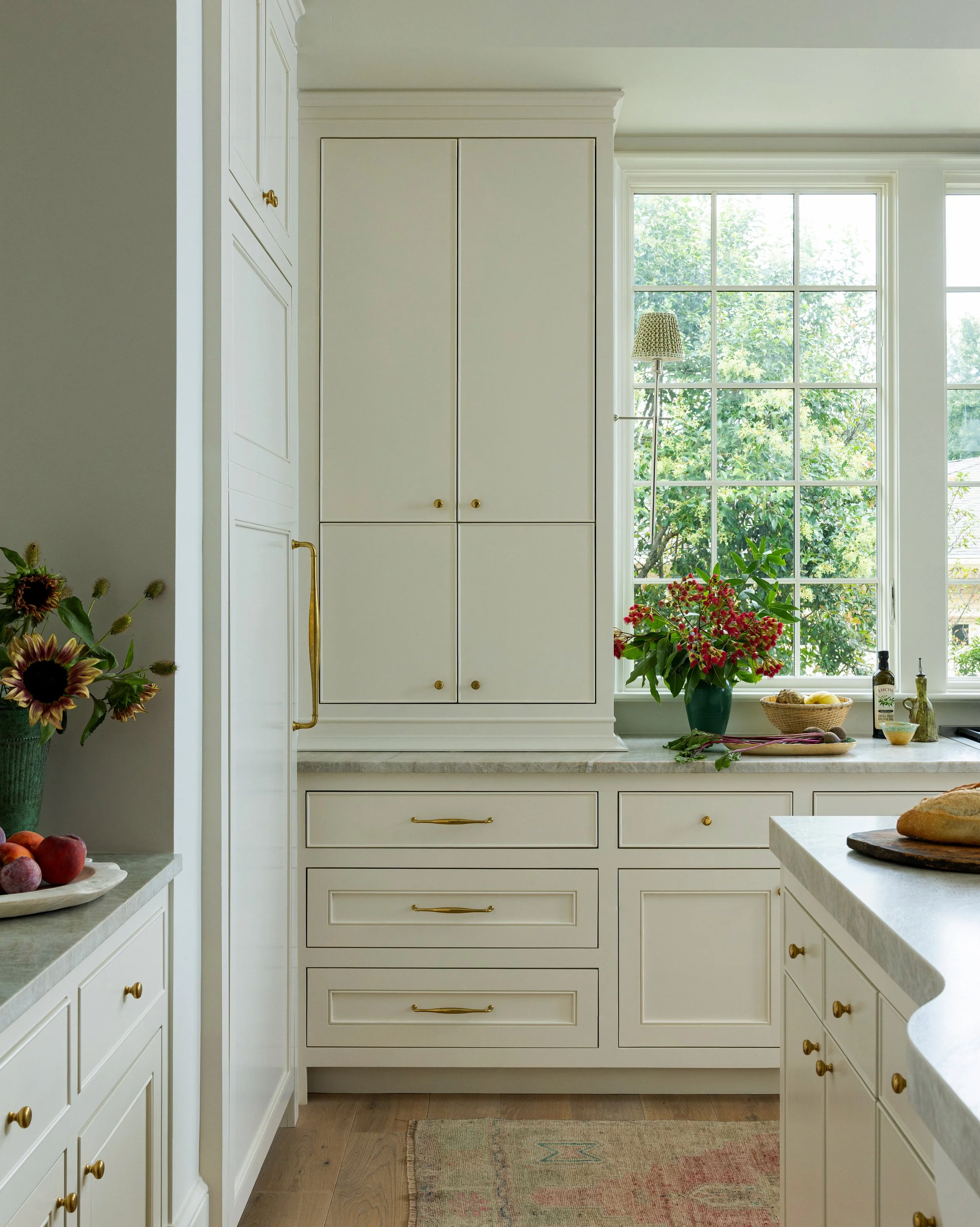 Bright kitchen with white cabinetry, gold handles, large window with greenery outside, countertop with a vase of pink flowers, loaf of bread, and various small kitchen items.