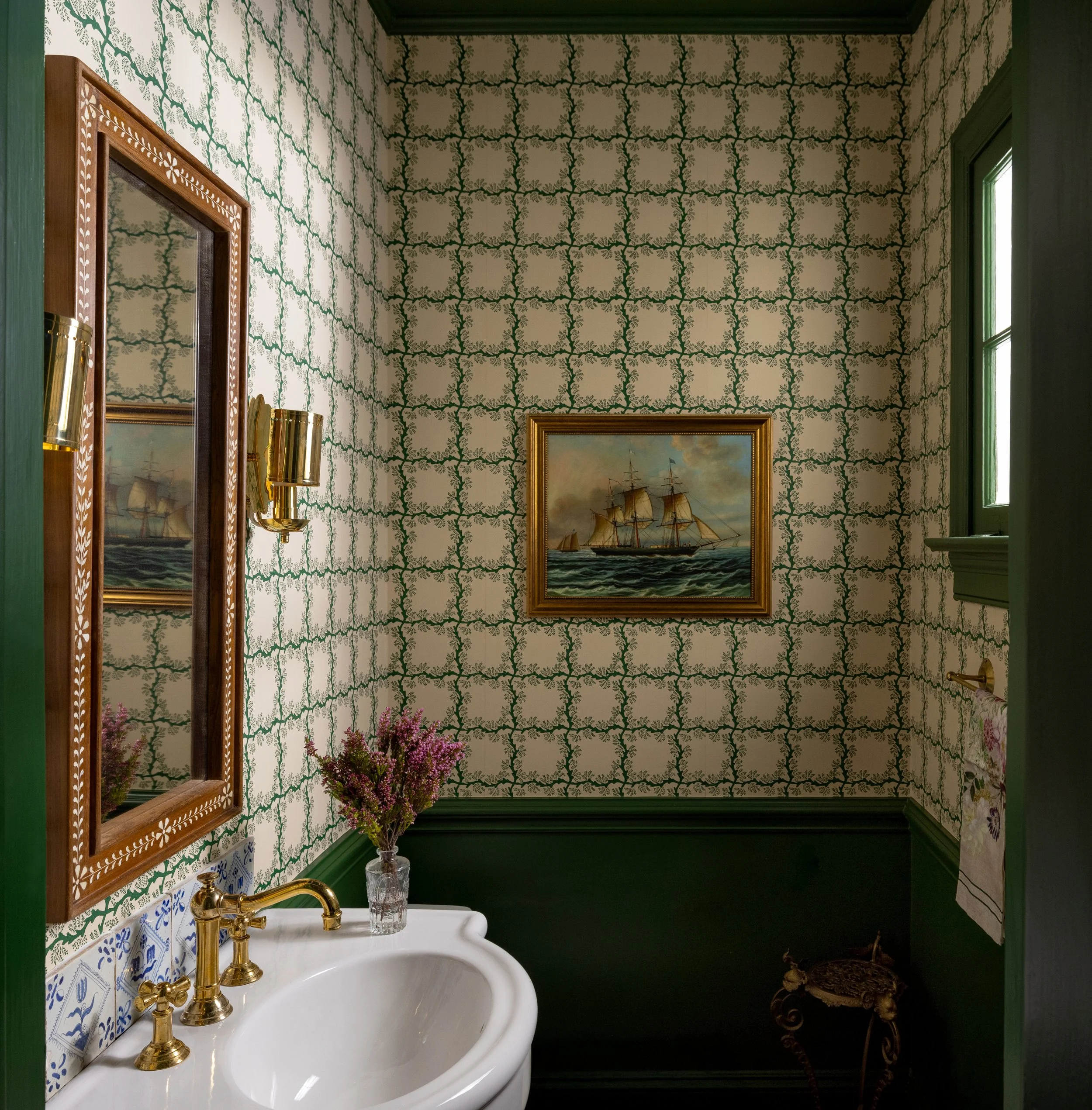 A vintage-style bathroom with patterned wallpaper, a white sink with gold fixtures, a mirror, a vase with purple flowers, a small painting of a sailing ship, green window trim, and a towel ring with a floral towel, with a small decorative table in th
