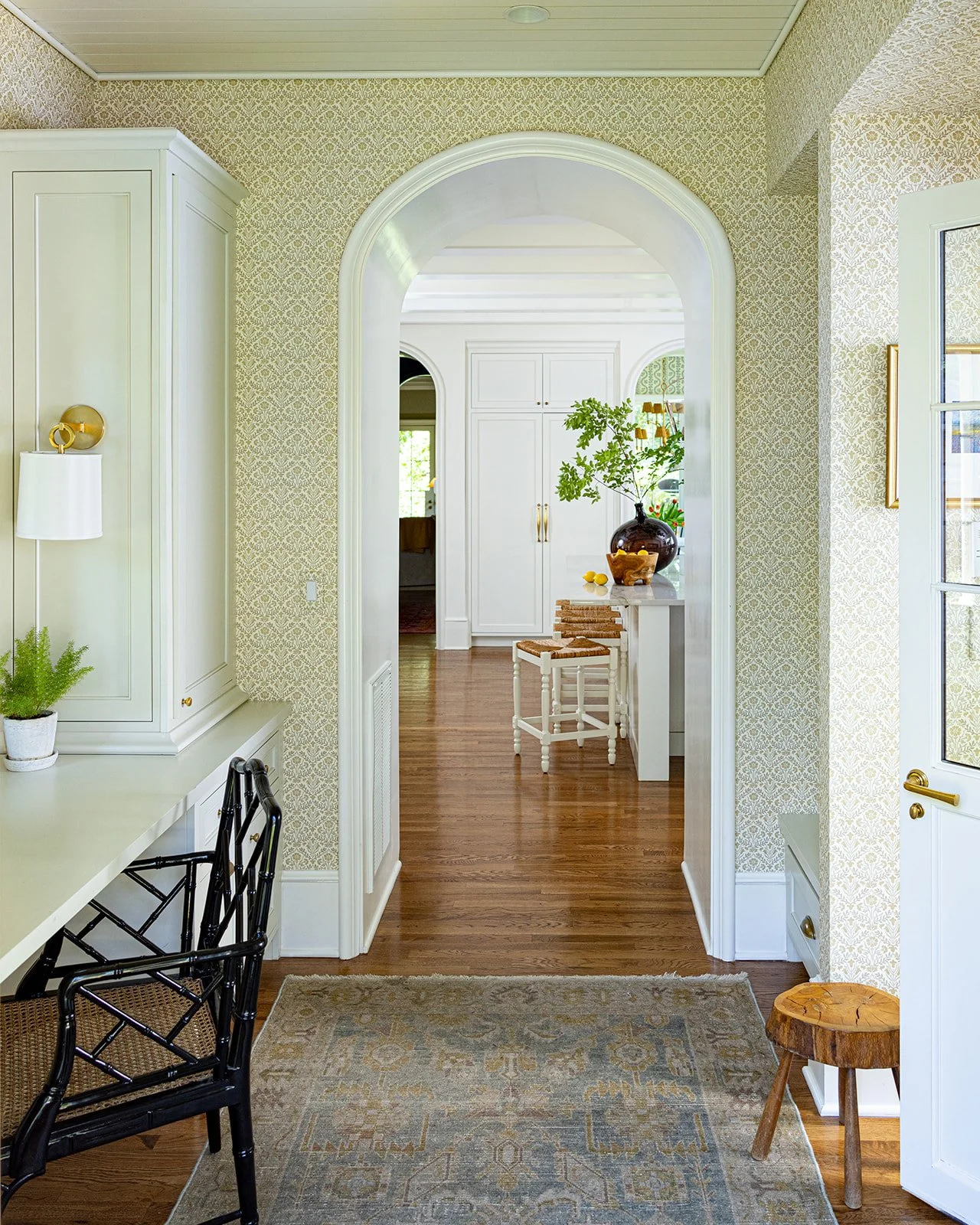 A cozy home interior featuring a hallway with wood flooring, patterned wallpaper, a black desk chair, a white desk with a small potted plant, and a doorway leading to a room with a white cabinet, a table with a large vase of greenery, and a wooden st