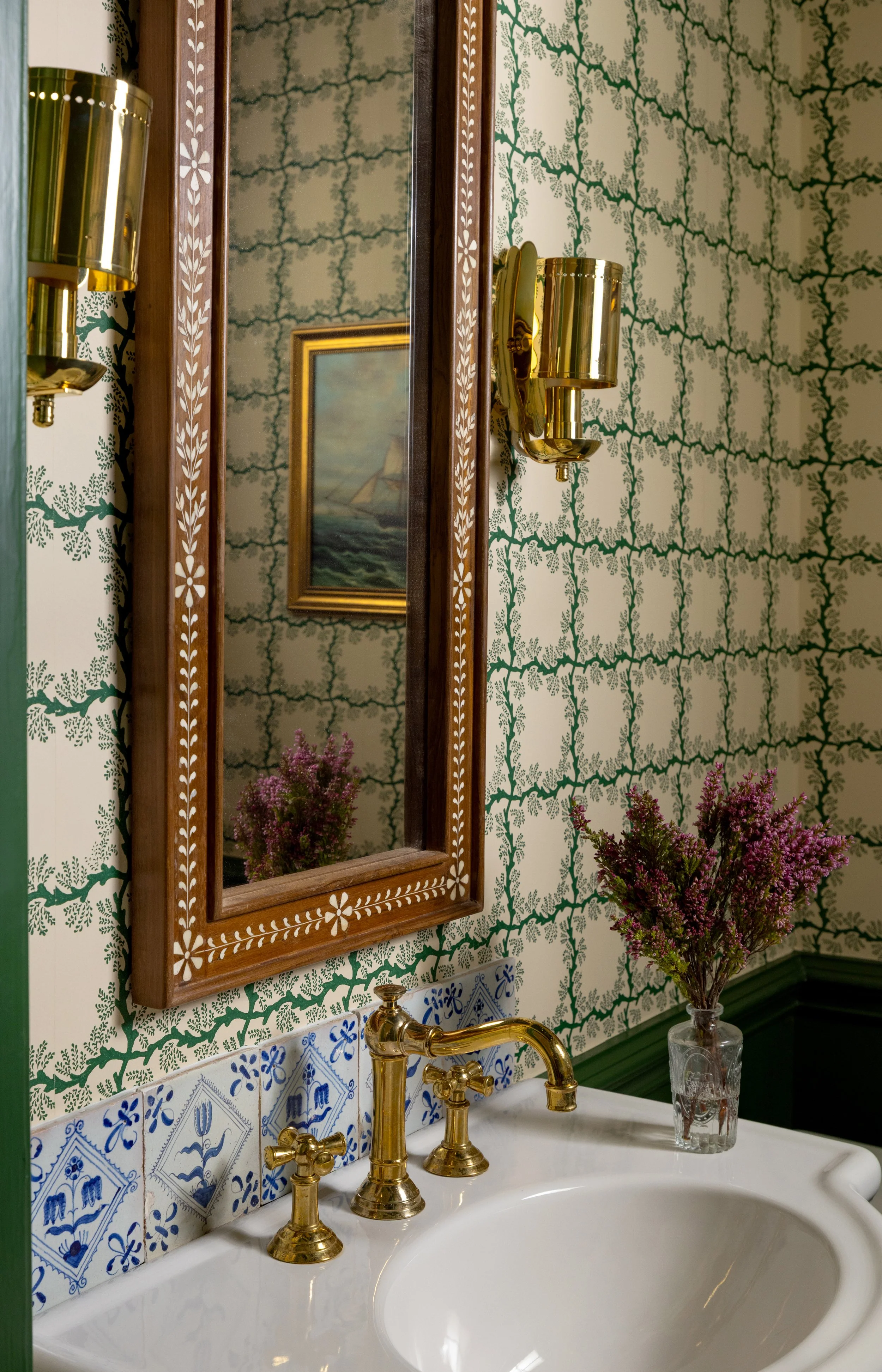 A bathroom sink with gold fixtures and a mirror surrounded by ornate wood framing. There is a small vase with purple flowers on the sink, and a wall lamp with a gold finish. The wallpaper features a green and white leaf motif, and the tiles beneath t