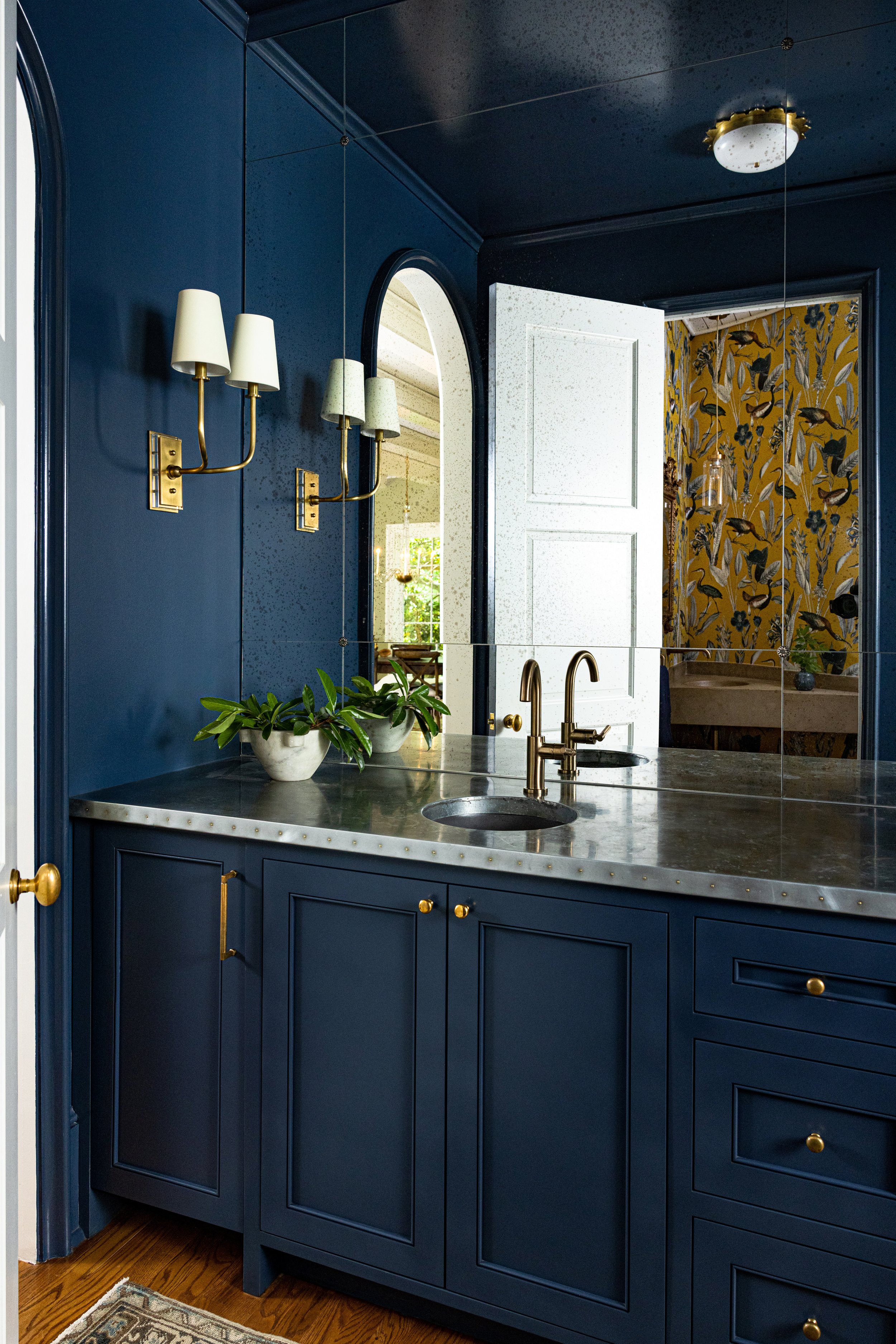 A bathroom with dark blue paneled walls, gold fixtures, a marble countertop, two plants in white pots, and a large mirror reflecting a doorway and part of a patterned wall.