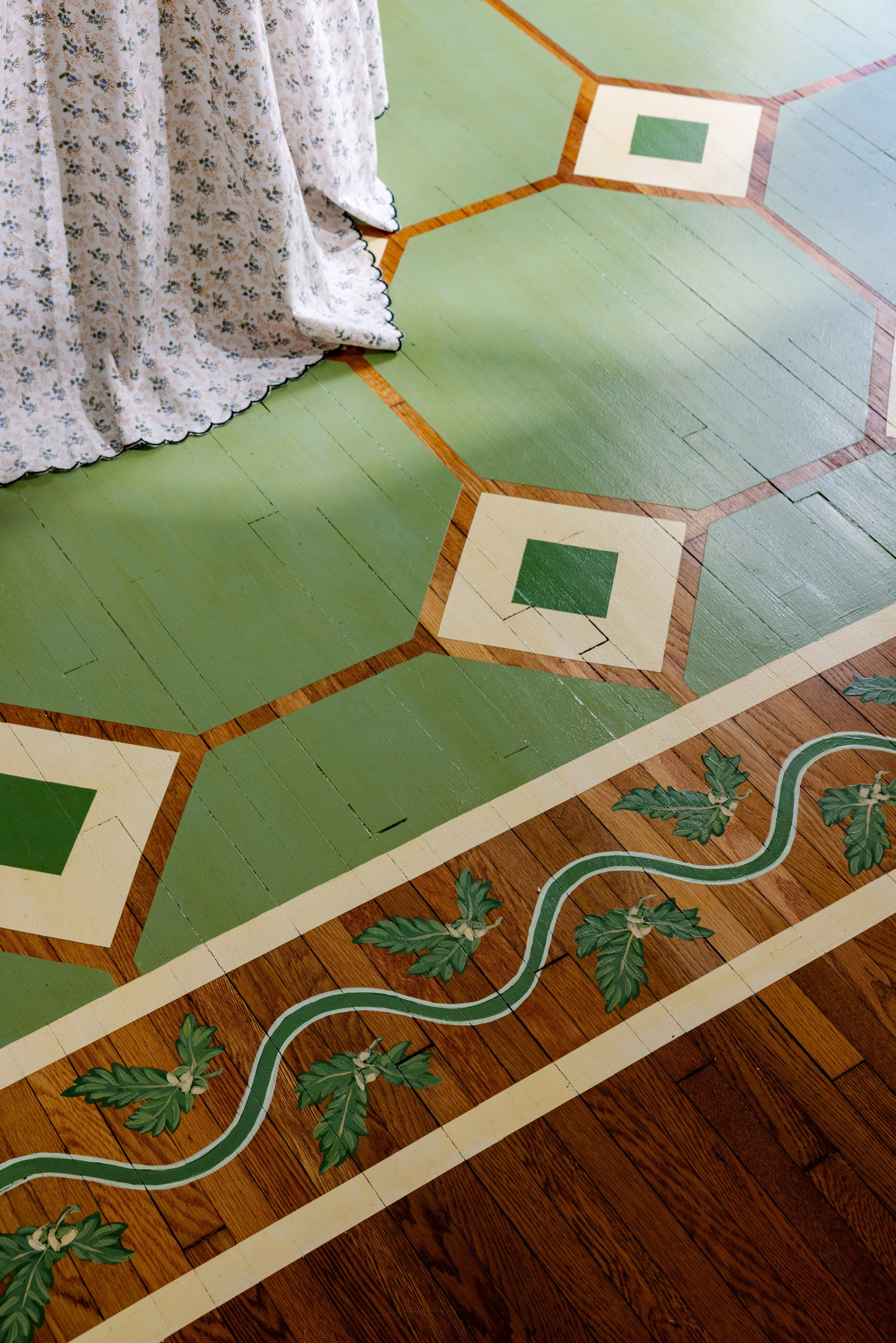 Vintage wooden floor with a geometric pattern of green and cream tiles, some with cracks, and a border decorated with vines and leaves.