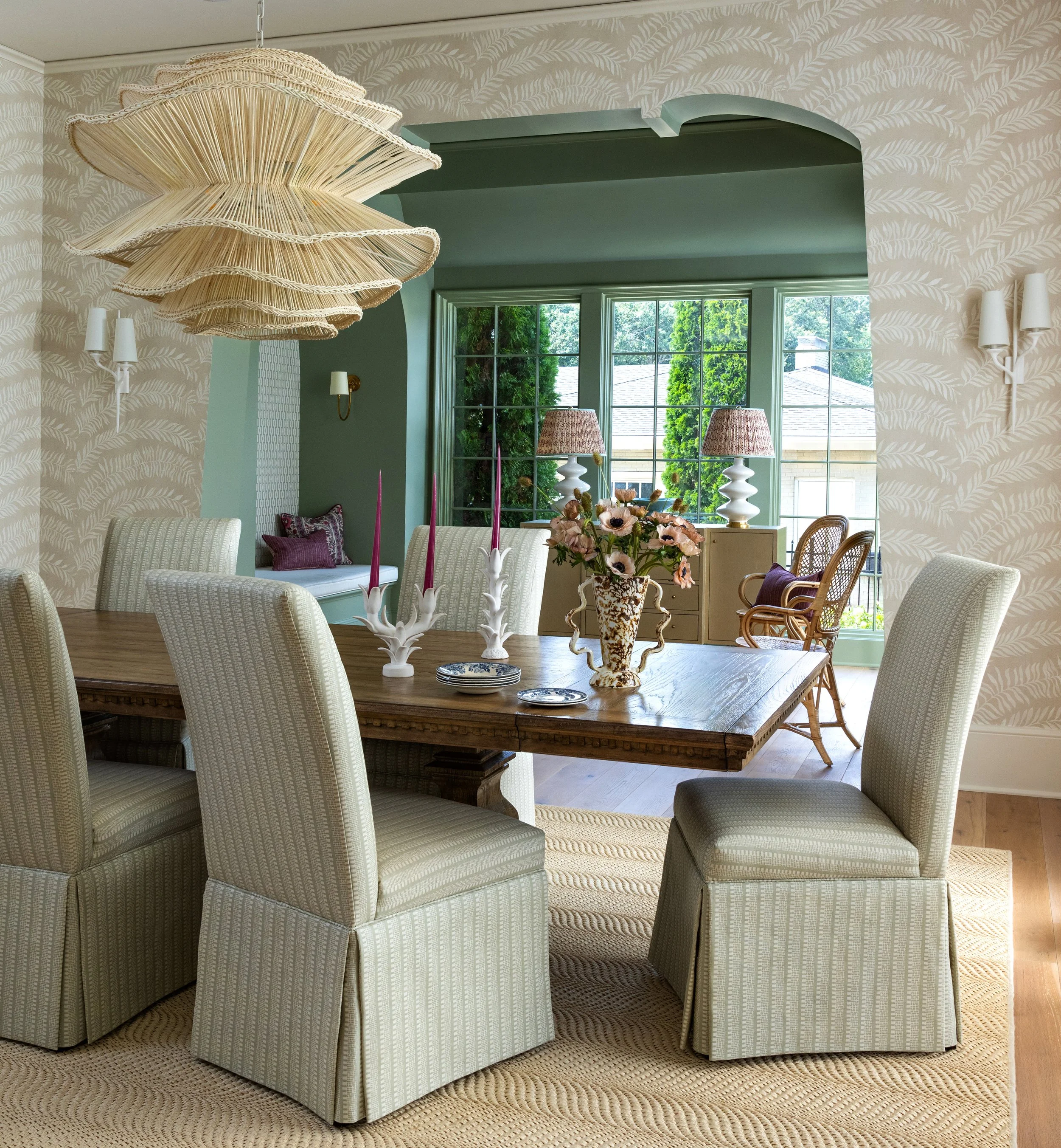 A dining room with a wooden table surrounded by six upholstered cream-colored chairs. On the table are candle holders with pink candles and a vase with pink flowers. The room has wallpaper with a feather pattern, a large hanging light fixture made of