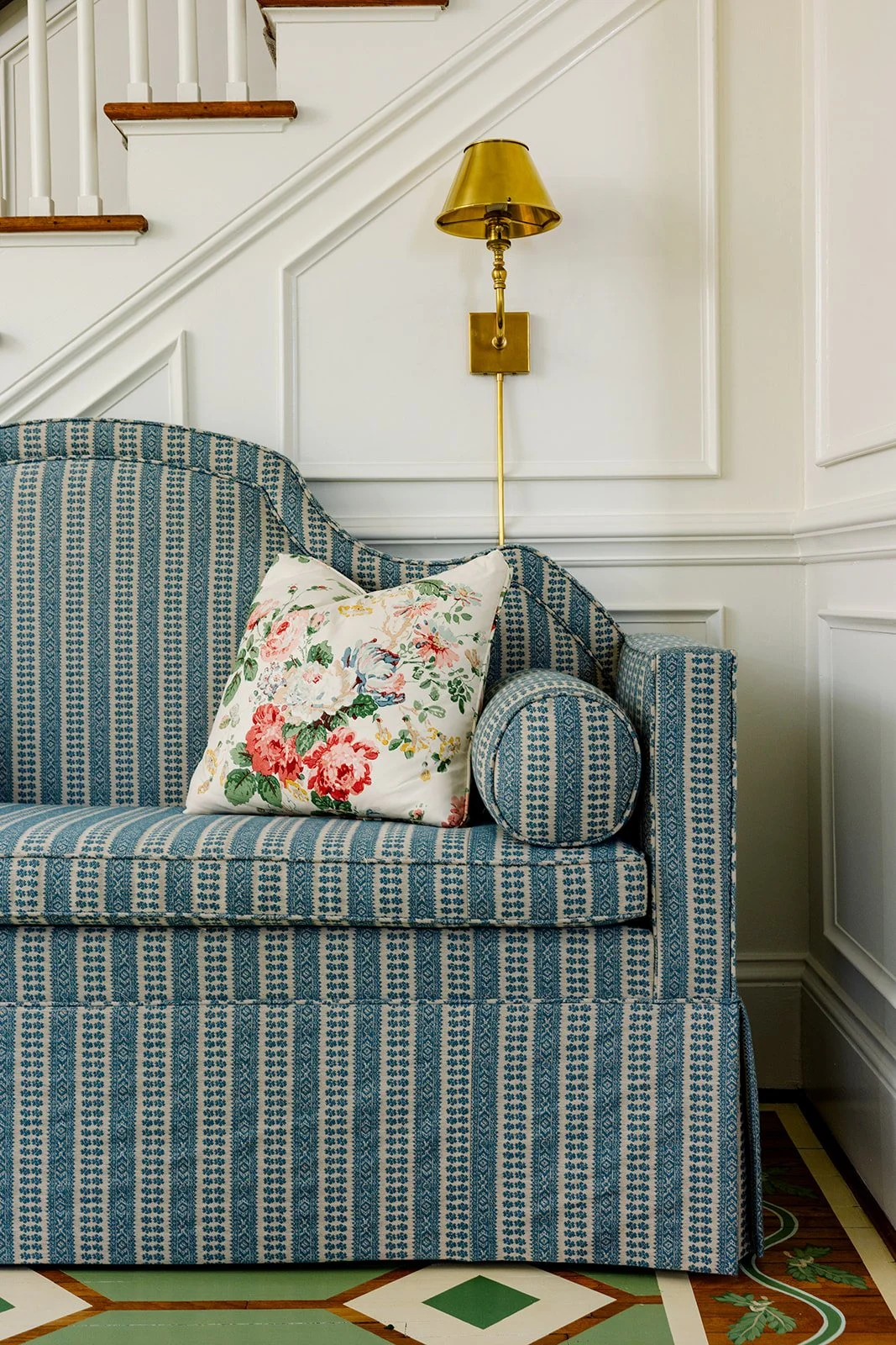 A blue patterned loveseat with a floral pillow, a gold wall sconce with a shade, and white paneled walls with wood trim.