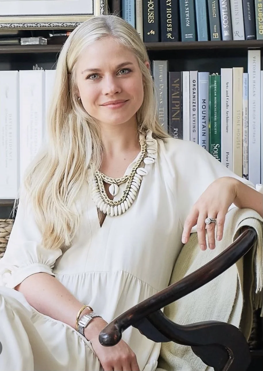 A woman with long blonde hair seated next to a bookshelf filled with various books, wearing a cream-colored dress and layered necklaces, resting her arm on the back of a chair.