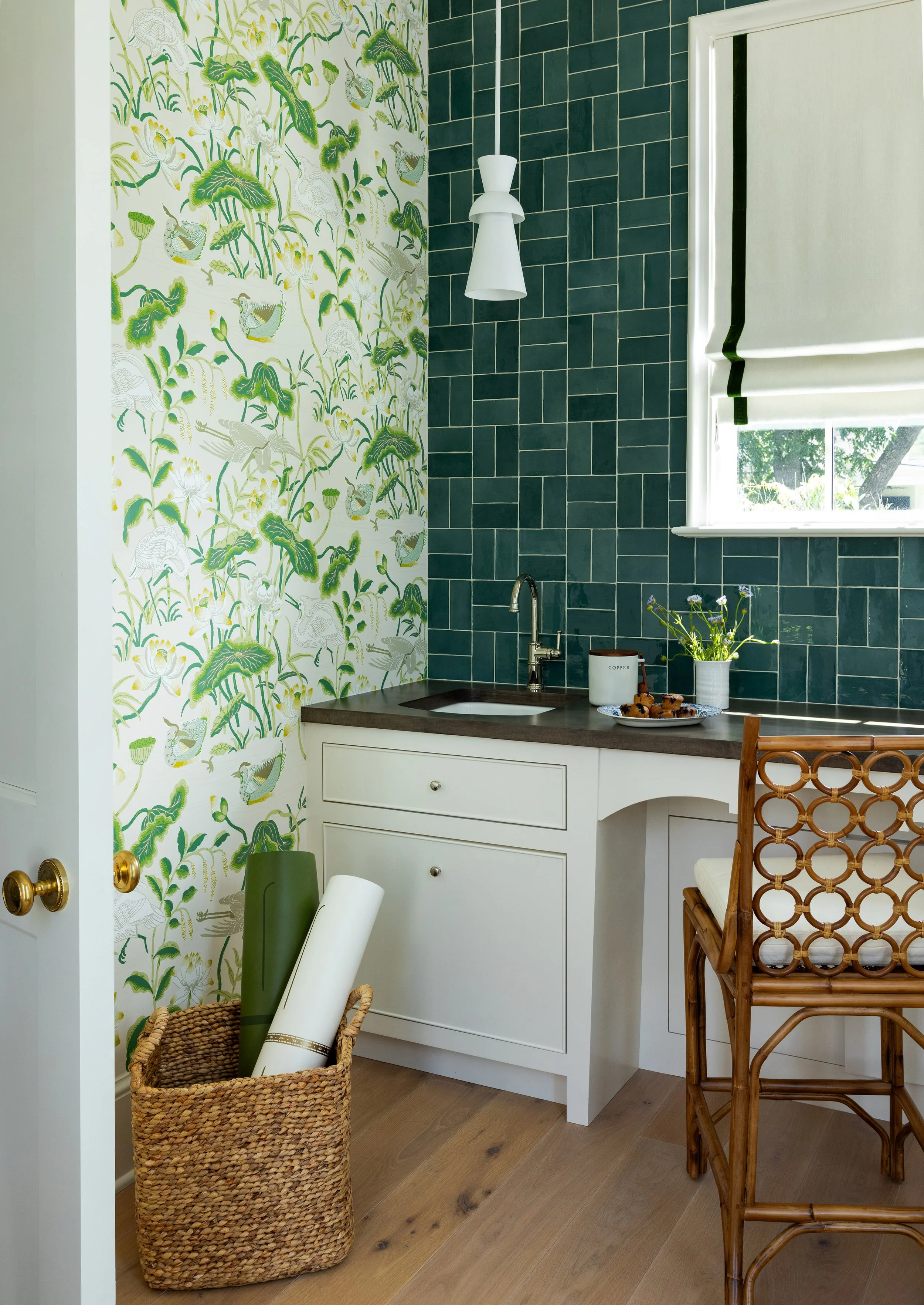 Small kitchen with green tiled backsplash, a white cabinet with a dark countertop, a small sink, a window with a blind, and decorative items including a vase with flowers, a cup labeled 'coffee,' and some cookies on a plate. There is a woven basket w