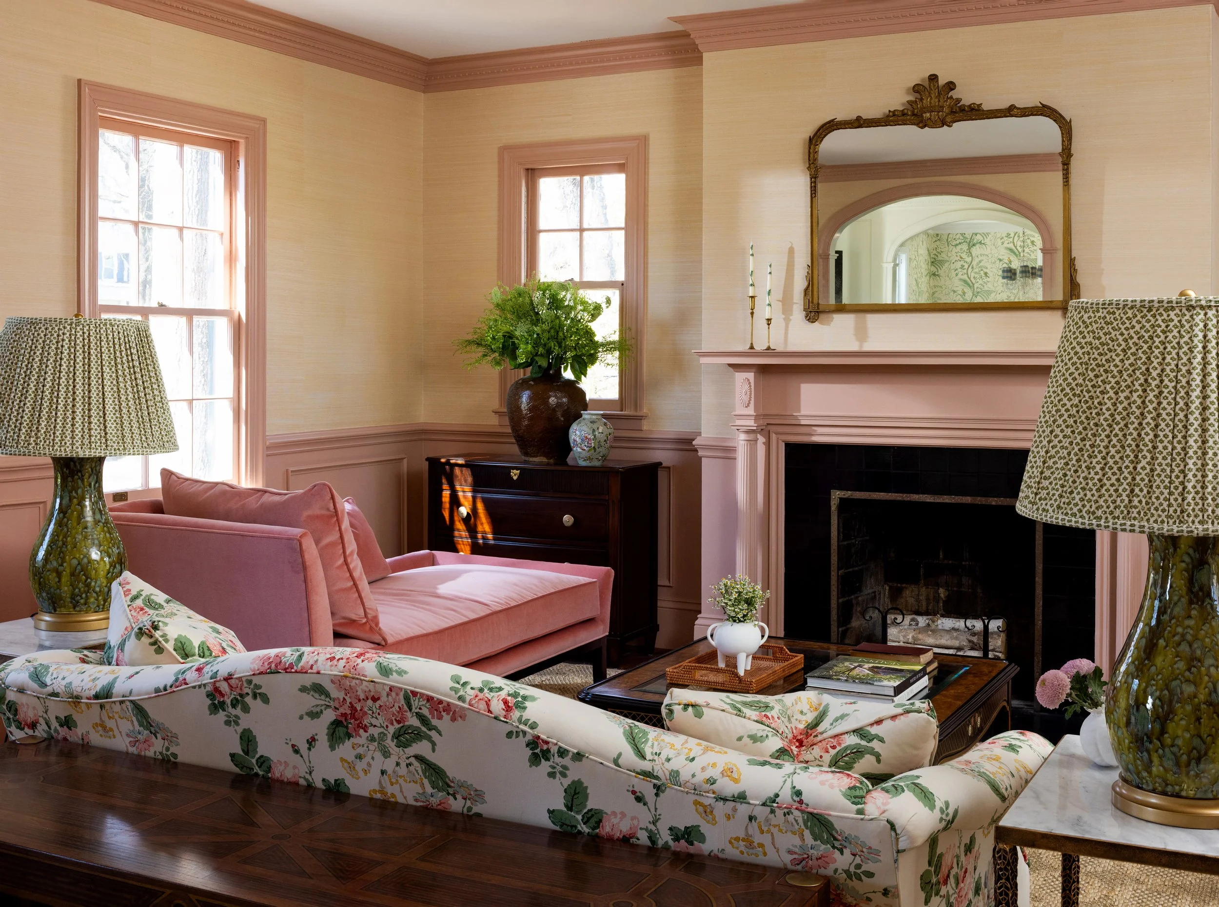Living room with pink walls, floral and pink furniture, a fireplace with a gold-framed mirror above, and a vase with green foliage on the window sill.