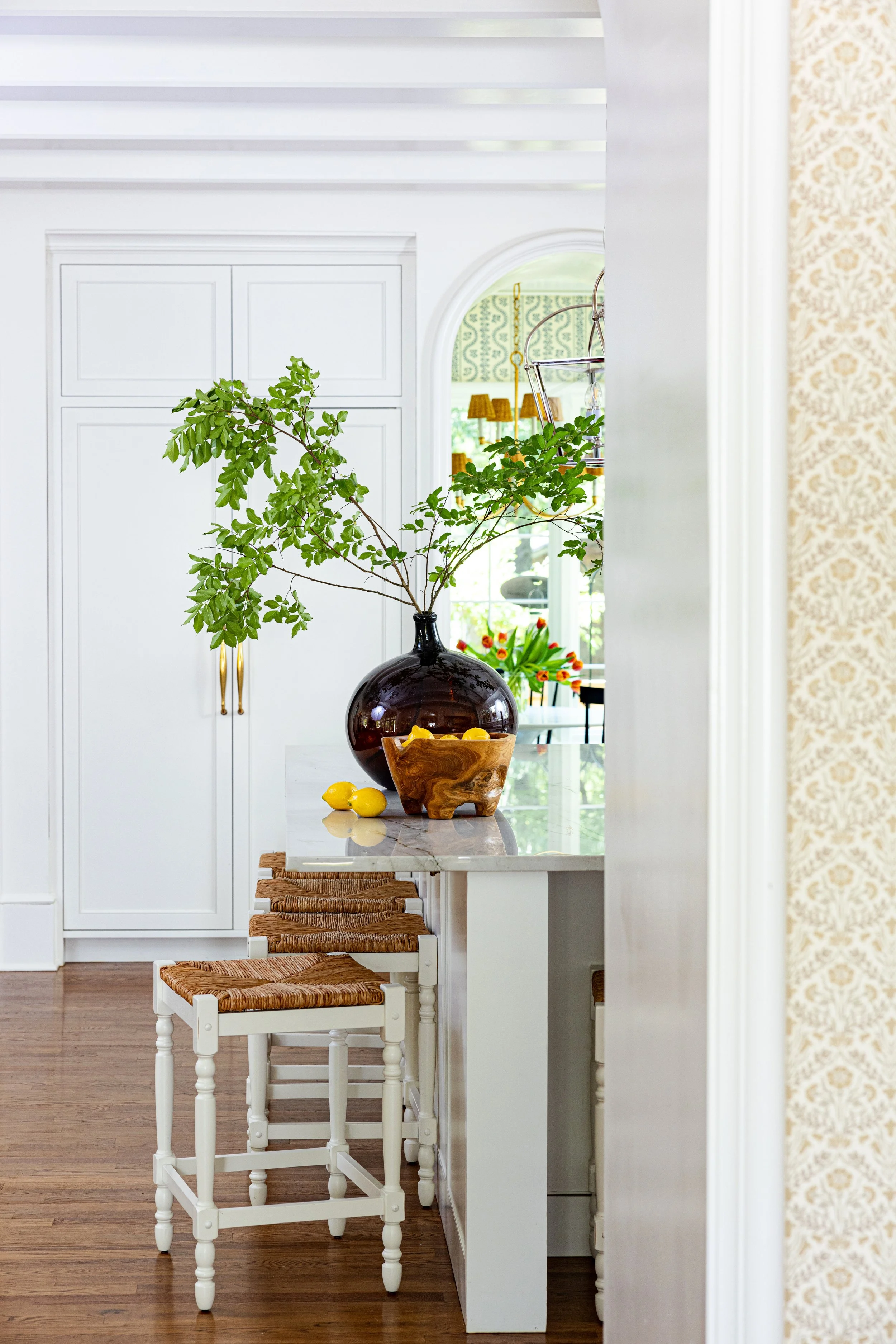 A white kitchen island with a black vase containing green leafy branches, a wooden bowl with lemons, and three woven stool seats underneath a marble countertop. White cabinets and a window with floral arrangements are visible in the background.