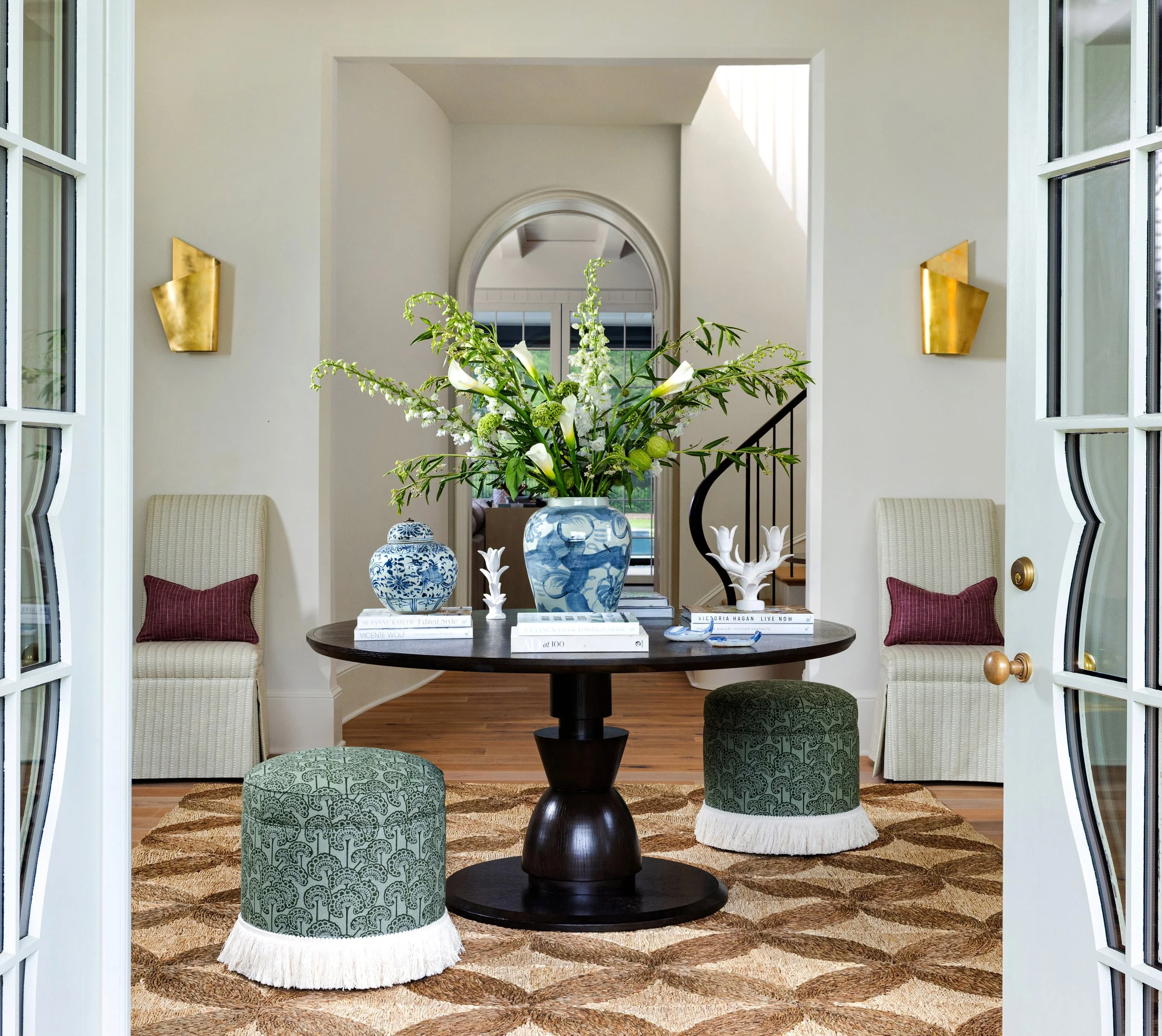 An entryway with French doors opening into a circular dining area. The room features a dark round table with a large floral arrangement, decorative books, and ceramics. Two upholstered green ottomans with white fringe are in the foreground, and two c