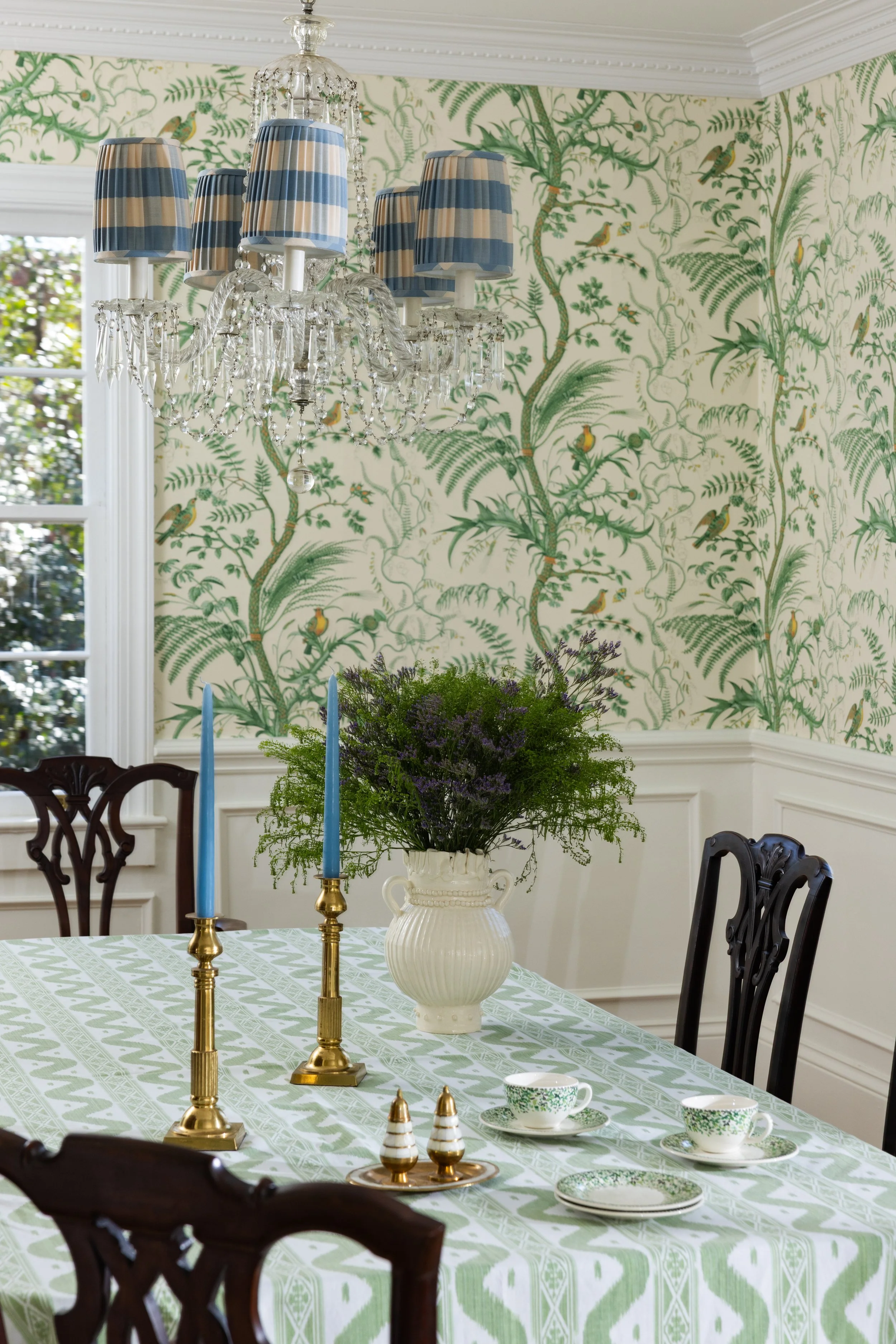 Dining room with a table set for tea, featuring a green patterned tablecloth, white teacups with green patterns, and gold candlesticks with blue candles. A white vase with purple and green flowers sits in the center, and a crystal chandelier with blu