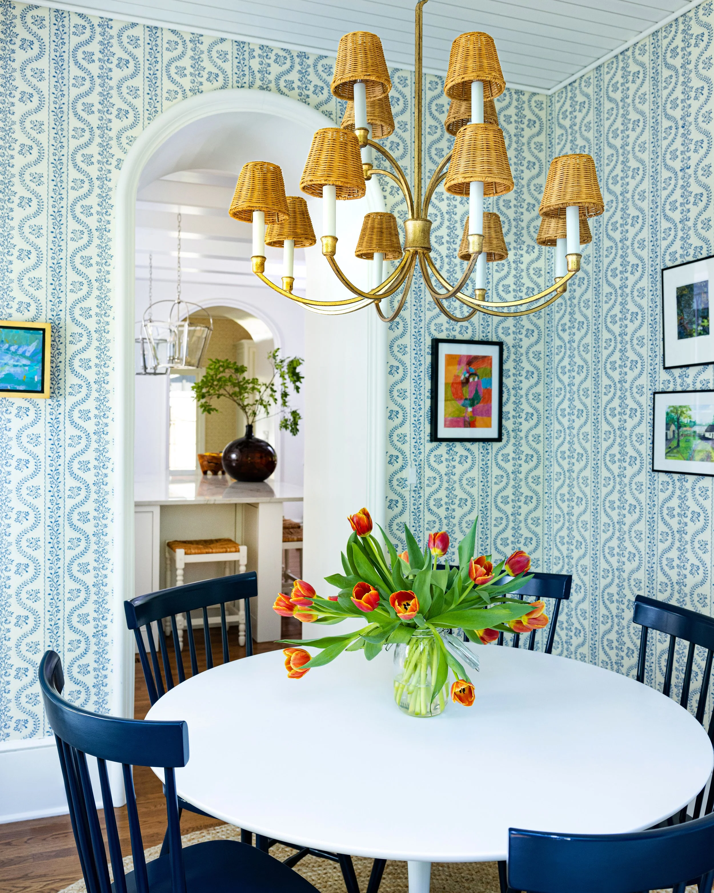 A dining room featuring a white round table with a glass vase of red and yellow tulips, surrounded by black chairs. A wicker chandelier hangs above, and the walls are covered in blue and white patterned wallpaper. There are framed pictures on the wal