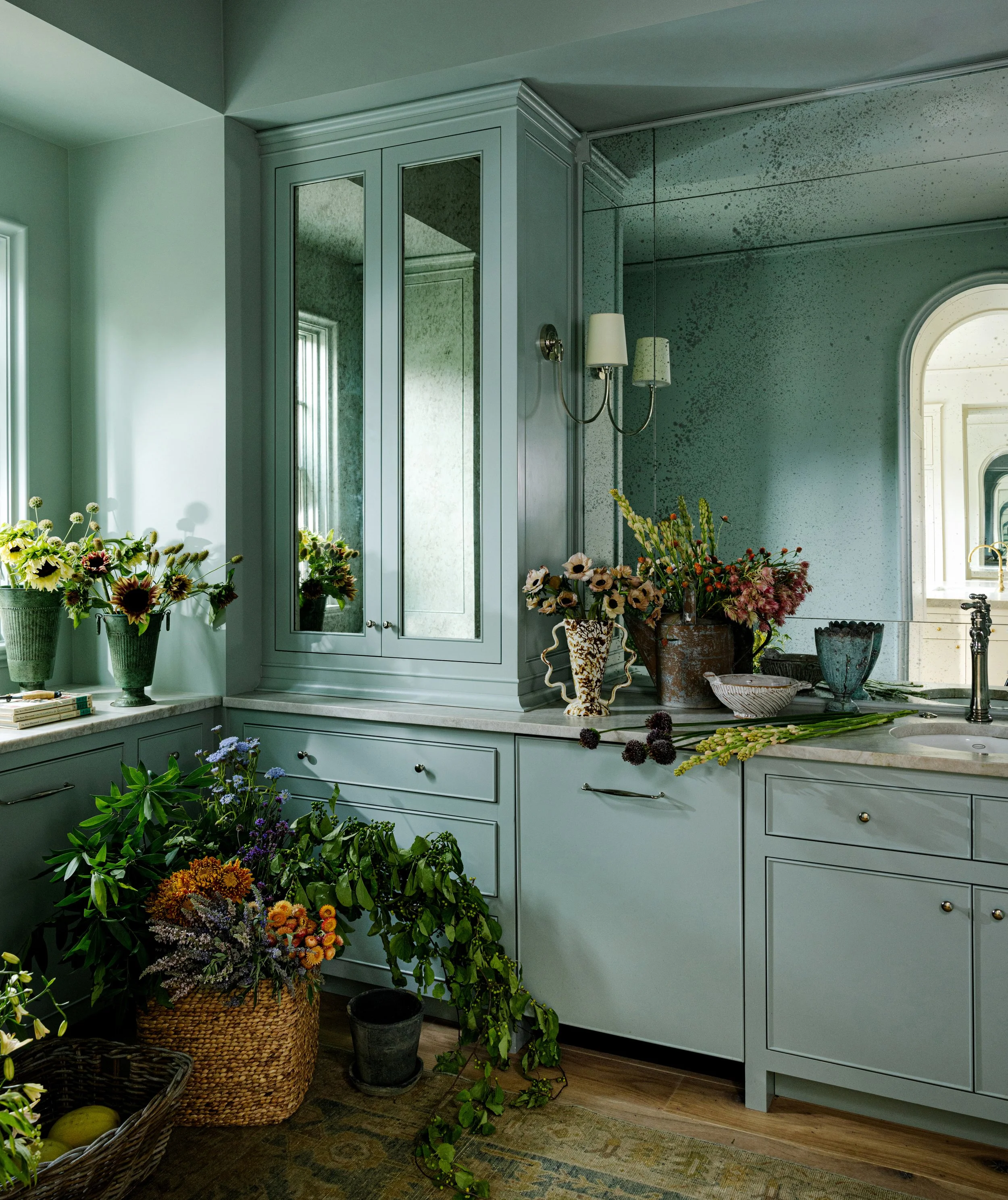 A kitchen corner with pastel green cabinets and drawers, decorated with various potted plants and flowers in vases, a mirror wall, a window letting in natural light, and a wooden floor with a rug.