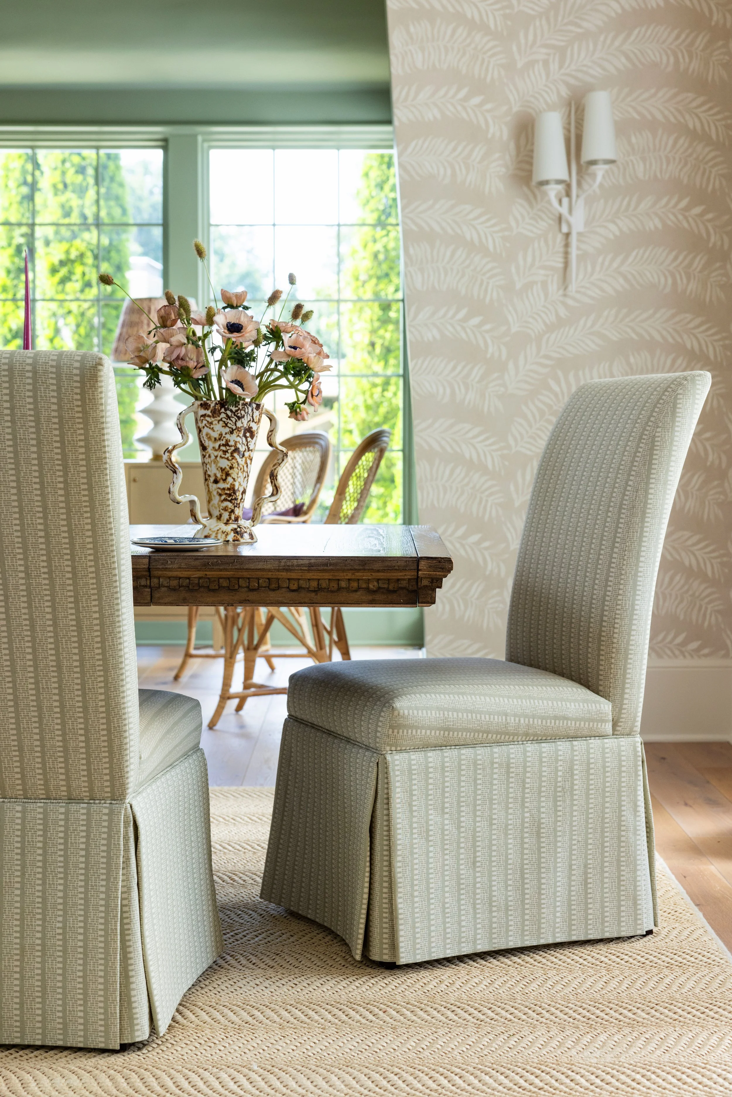 A dining room with upholstered chairs, a wooden table, a floral centerpiece in a decorative vase, and large windows showing greenery outside.