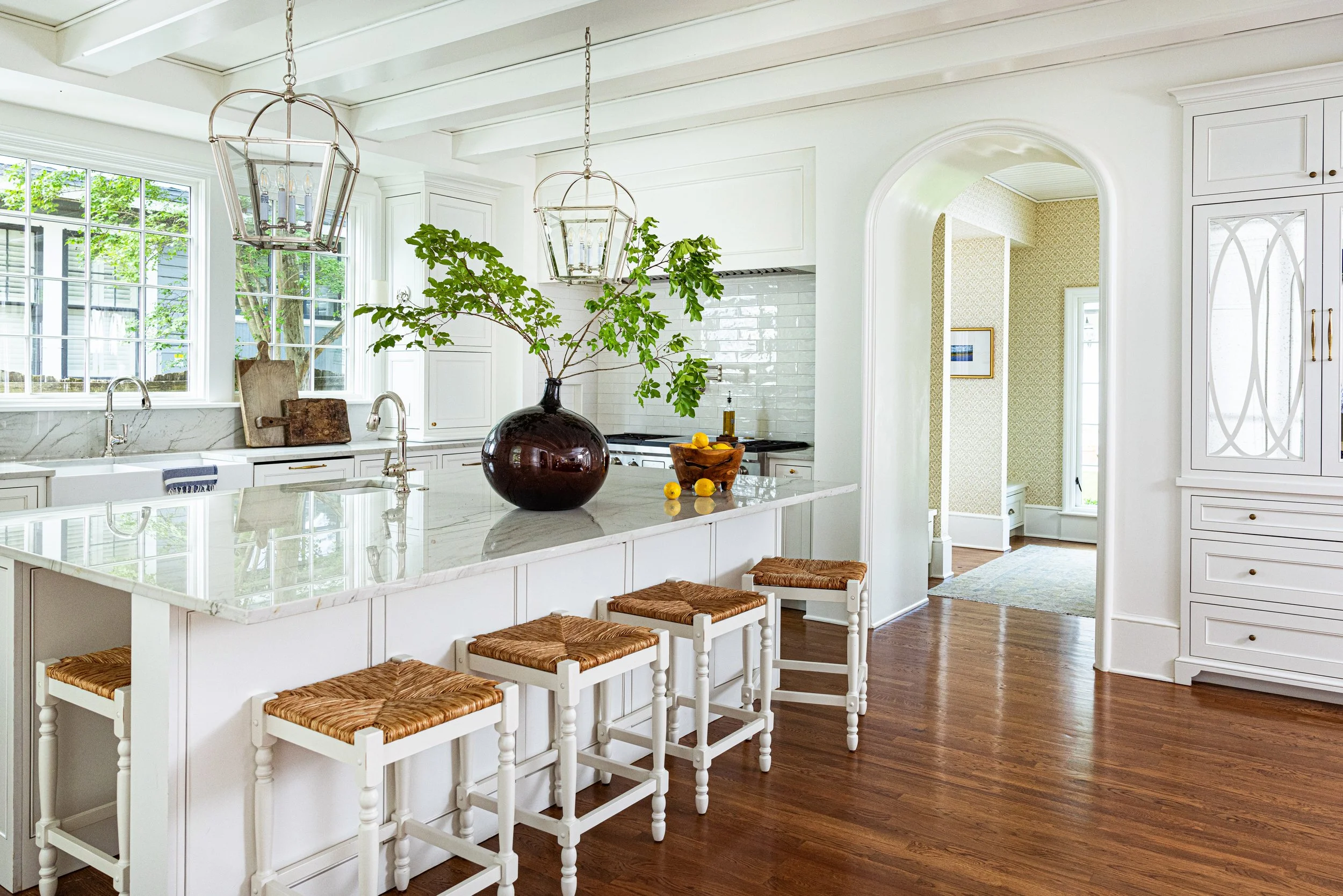 Bright white kitchen with a marble island, four wicker stools, and large window showing greenery outside. The kitchen features white cabinets, a green plant in a dark vase on the island, and two pendant lights hanging from the ceiling.