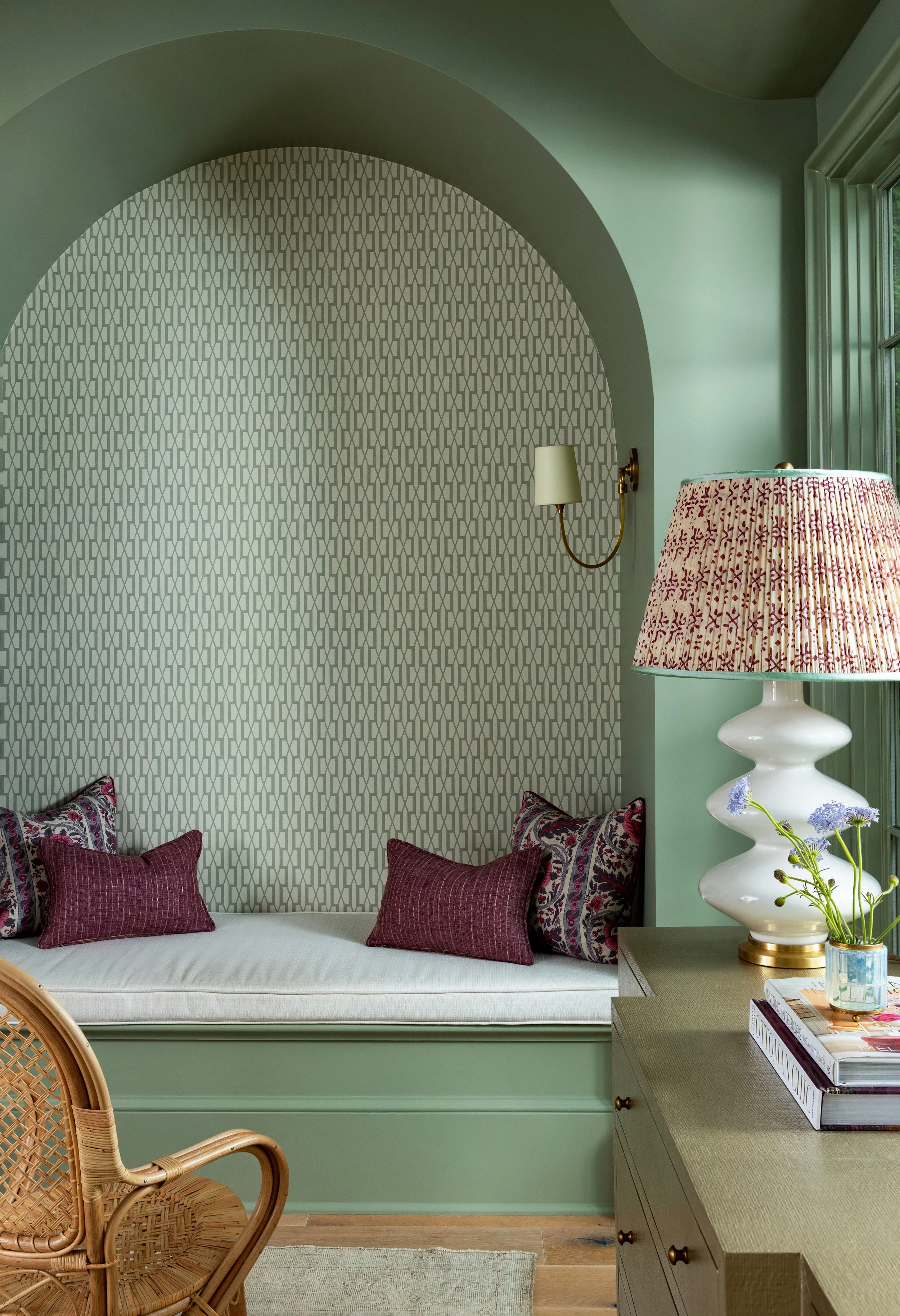 A cozy window seat with patterned pillows, a lamp, and a side table with flowers in a room with green walls and a patterned accent wall.