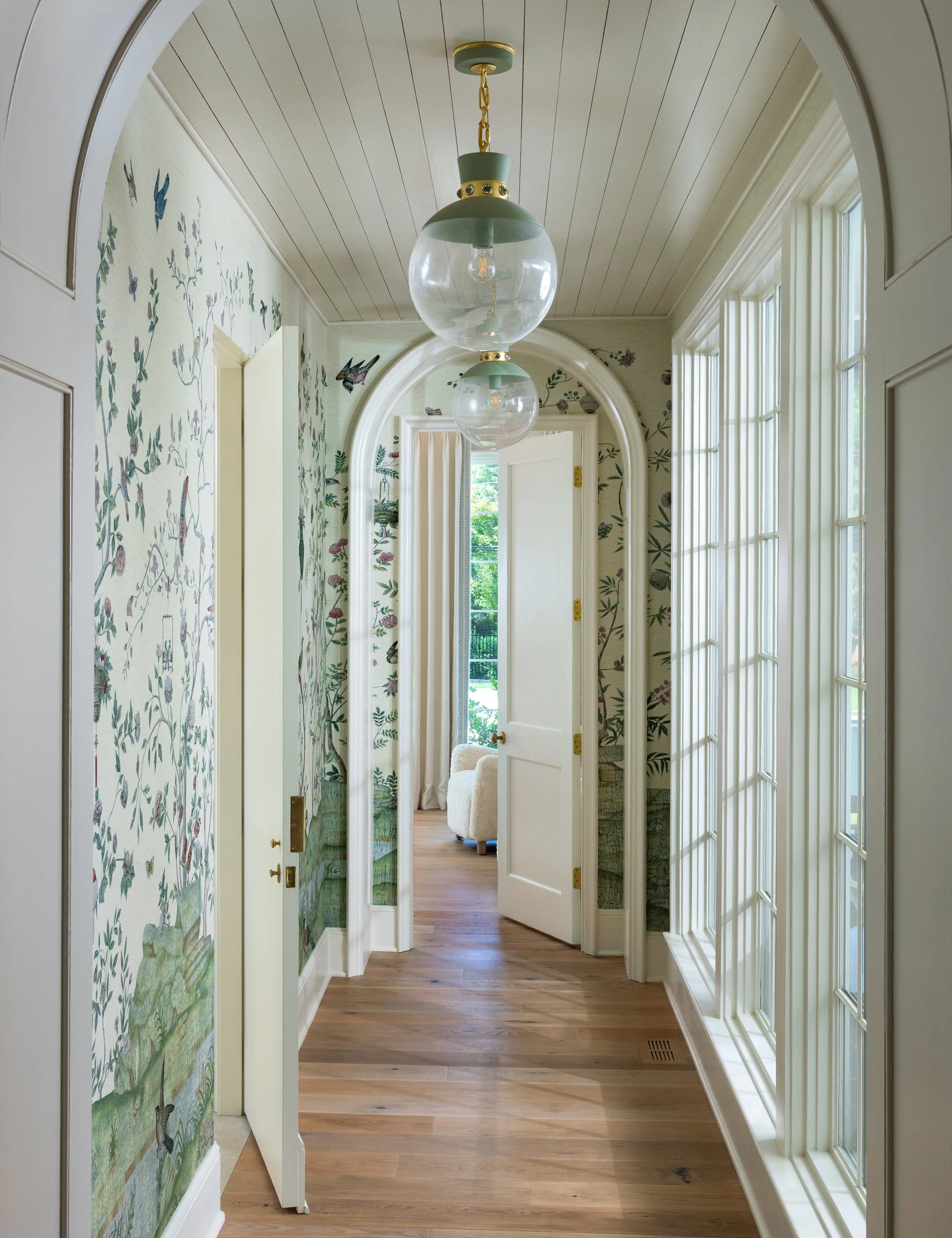 A bright hallway with wooden flooring, floral wallpaper, large windows on the right, an arched doorway, and a hanging glass globe light fixture.